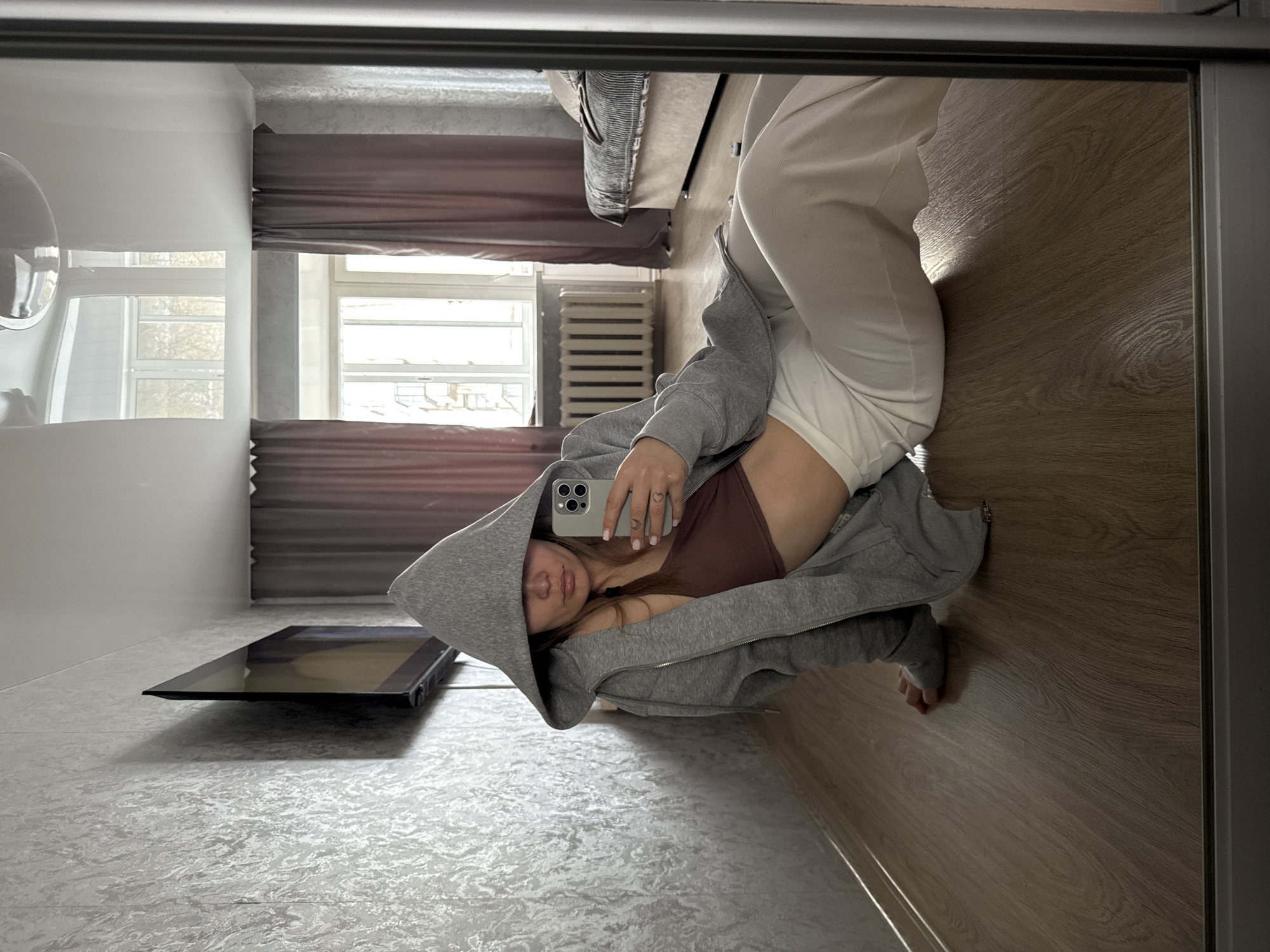 A woman is taking a selfie in a mirror. She is wearing a grey hoodie, a brown top, and white bottoms. The room features a window with brown curtains, a radiator, and a TV on a textured surface. The floor is a mix of textured and wood-grain surfaces. The reflection suggests the location might be in an apartment or home in Chaikovsky, Russia.