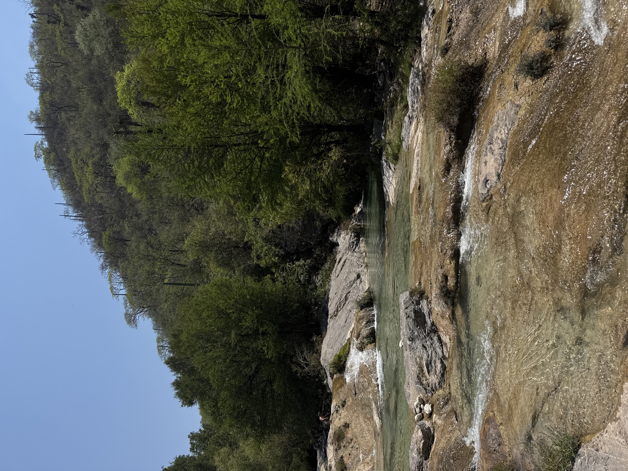 The image depicts a waterfall and river in a natural setting, possibly located near San Miguel Atlapulco, Mexico. A steep, tree-covered hillside rises on the left, transitioning to a rocky cliff and water cascading over the falls on the right. The river flows in the foreground, with visible rocks and clear water. A few people can be seen at the base of the falls.