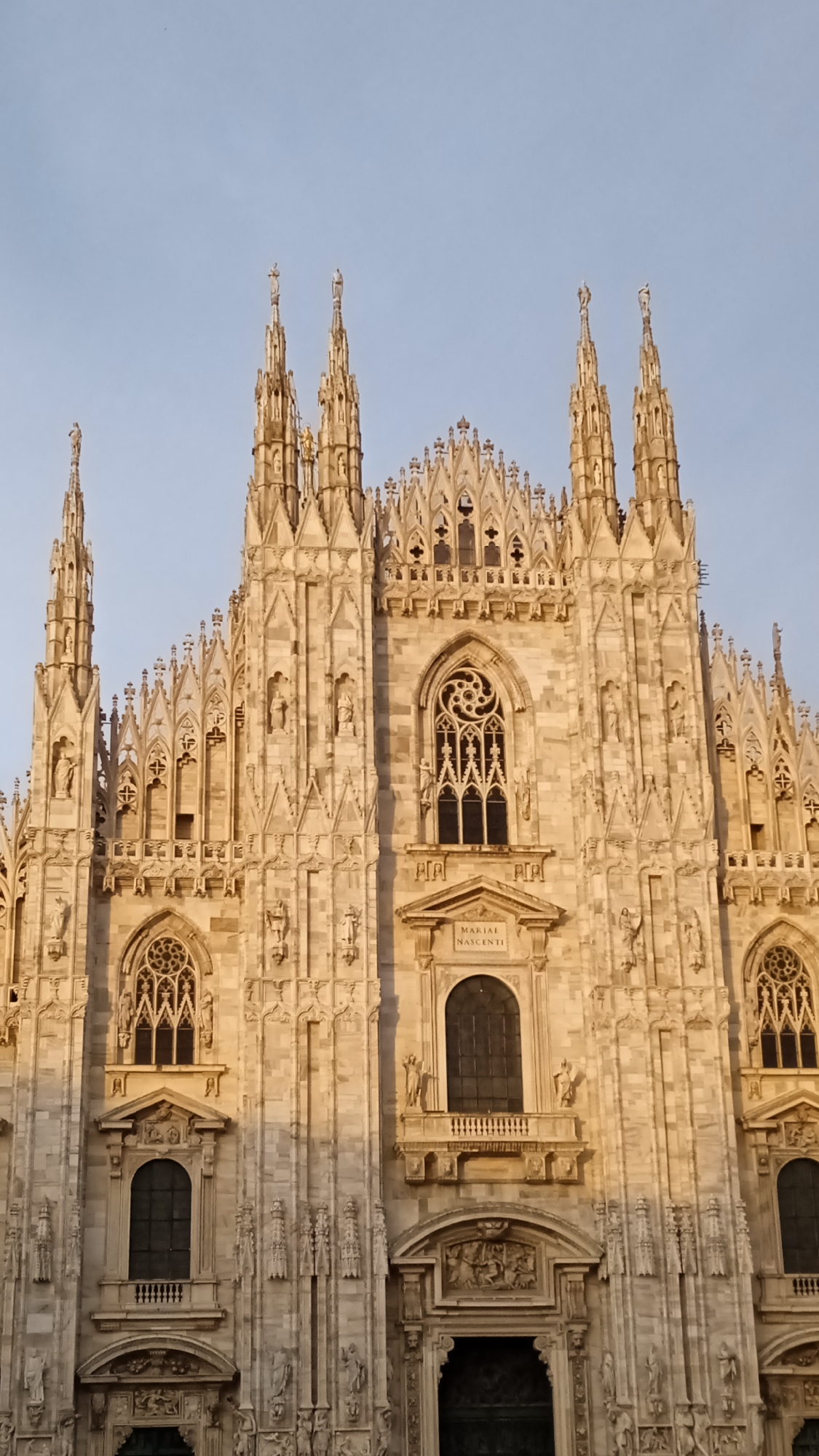 The image shows the facade of the Milan Cathedral, also known as the Duomo di Milano, in Milan, Italy. The cathedral is made of white marble and is characterized by its intricate Gothic architecture, featuring numerous spires, statues, and decorative elements. The photograph is taken from a low angle, looking up at the cathedral. The sky above is a clear, light blue, suggesting it is daytime. The sunlight illuminates the facade, casting shadows and highlighting the detailed stonework. The cathedral's grand entrance is visible at the bottom center of the image, with ornate carvings above the doorway. The architectural style is highly detailed and ornate, typical of Gothic cathedrals. There are no people visible in the image.