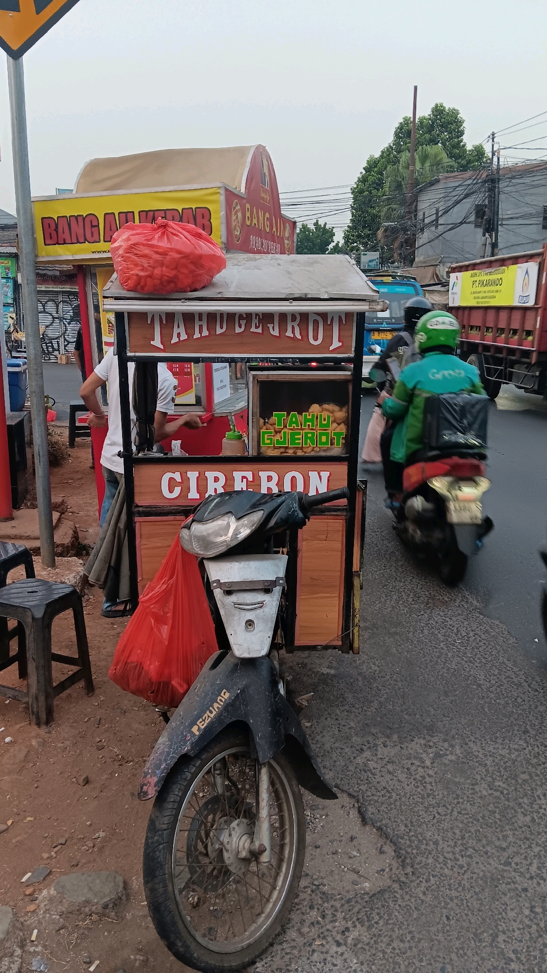 The image captures a street scene in South Jakarta, Indonesia, focusing on a food cart selling "Tahu Gejrot Cirebon". The cart, constructed from wood and metal, is adorned with banners displaying "BANG AJI KEBAB" and "TAHU GEJROT CIREBON". A large red plastic bag, possibly containing ingredients, is prominently placed on top of the cart. A person, partially obscured, stands behind the cart, seemingly attending to customers. In the foreground, a black and silver motorcycle is parked, its front wheel and fender clearly visible. The background reveals typical urban elements: buildings, electrical wires, and passing vehicles, including a red truck with "PT. PIKARANDO" signage. A person wearing a green jacket and helmet rides a motorcycle in the background, suggesting active commerce and movement within the city. The ground is paved with asphalt and dirt.