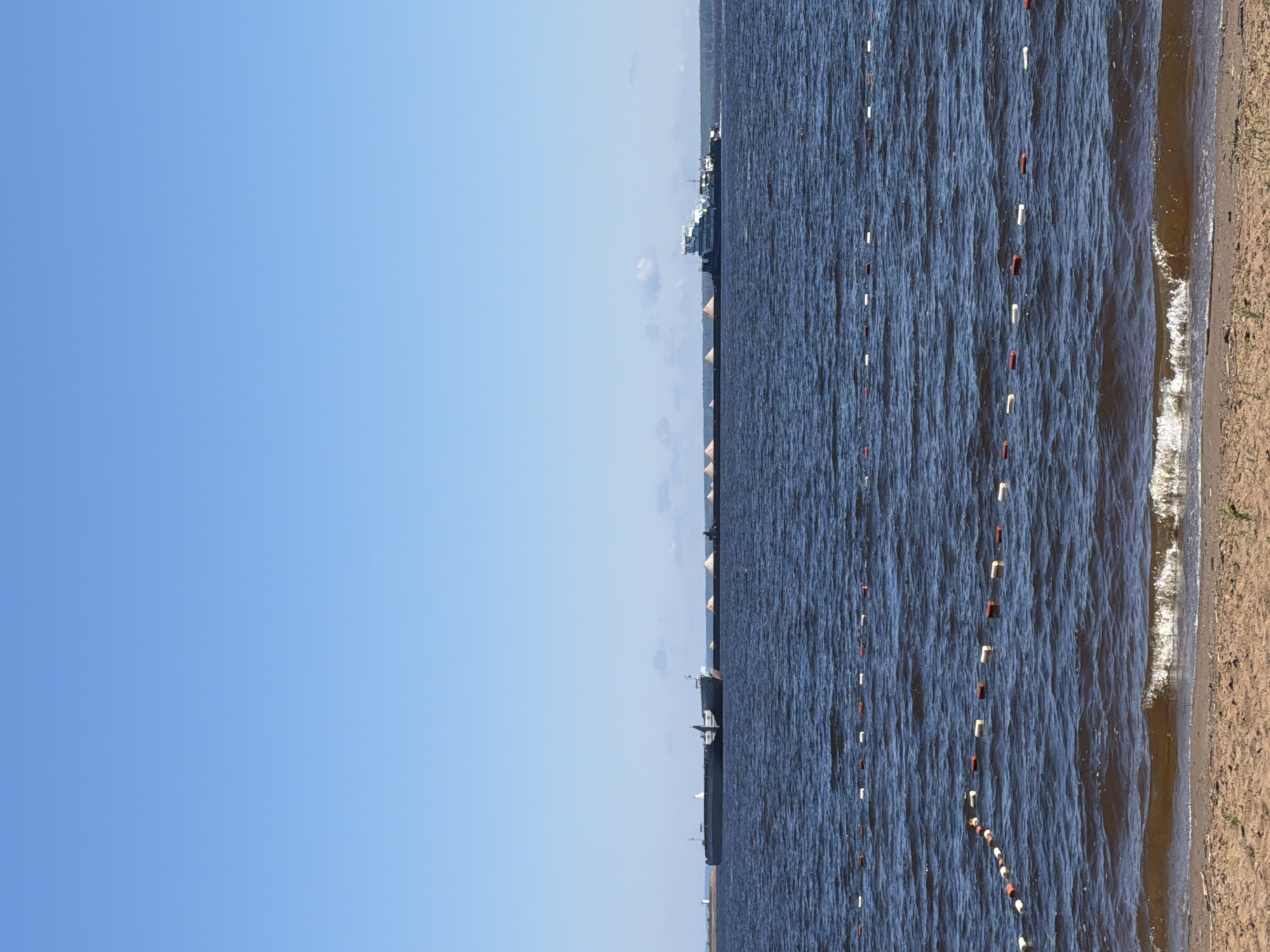 This image depicts a wide, calm body of water, likely a reservoir or a river, under a clear blue sky. In the distance, a long barge is being towed by a tugboat. The barge is laden with what appears to be sand or gravel, piled in conical mounds along its length. To the far left on the horizon, a white lighthouse is visible. Closer to the foreground, a series of buoys, alternating between red and white, mark a boundary in the water, indicating a swimming or restricted area. The shoreline in the immediate foreground is sandy with sparse vegetation, and small waves are lapping at the shore. The overall scene is serene and suggests a sunny day, possibly in a recreational area or a shipping channel. The location is identified as Chaikovsky, Russia, which is known for its location on the Kama River reservoir.