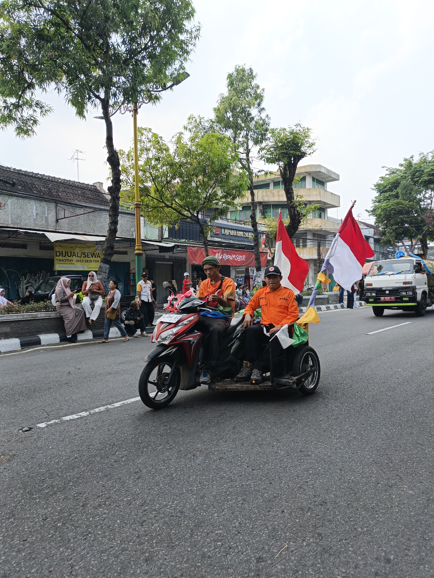 In the city of Bareng, Indonesia, a three-wheeled motorcycle with a sidecar is driving on a paved road. Two men are seated in the vehicle. The driver, wearing a green cap and an orange shirt, is holding onto the handlebars. Next to him, a passenger wearing an orange uniform and a black cap is also seated. Two Indonesian flags are attached to poles behind the sidecar. In the background, there are buildings, trees, and a white truck. Several people are visible on the sidewalk, some sitting and others standing. The sky is overcast.
