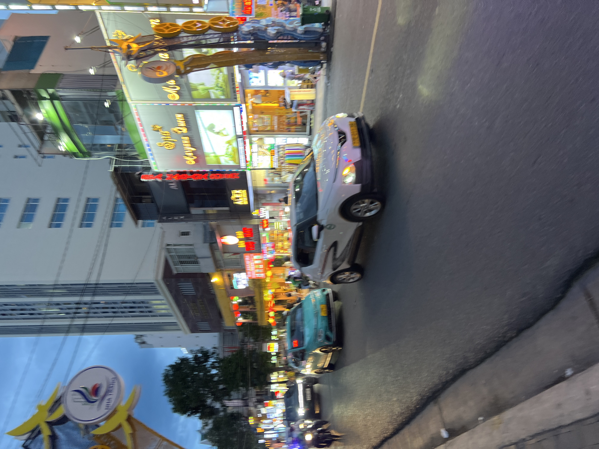 In Nha Trang, Vietnam, a street scene unfolds at dusk, captured with a slight motion blur. The image focuses on a busy road where several vehicles are present. In the foreground, a white car with its headlights illuminated is driving on the right side of the road. To its left, a turquoise taxi is also in motion. Further back, a black SUV is visible, and a motorcycle is seen on the far left. The buildings lining the street are brightly lit with neon signs. One prominent sign reads "Spa Maryana Queen". The sky above is a dusky blue, and power lines crisscross the frame. The architecture of the buildings varies, with a tall, modern high-rise visible in the background. The overall impression is of a vibrant urban environment at the transition from day to night.