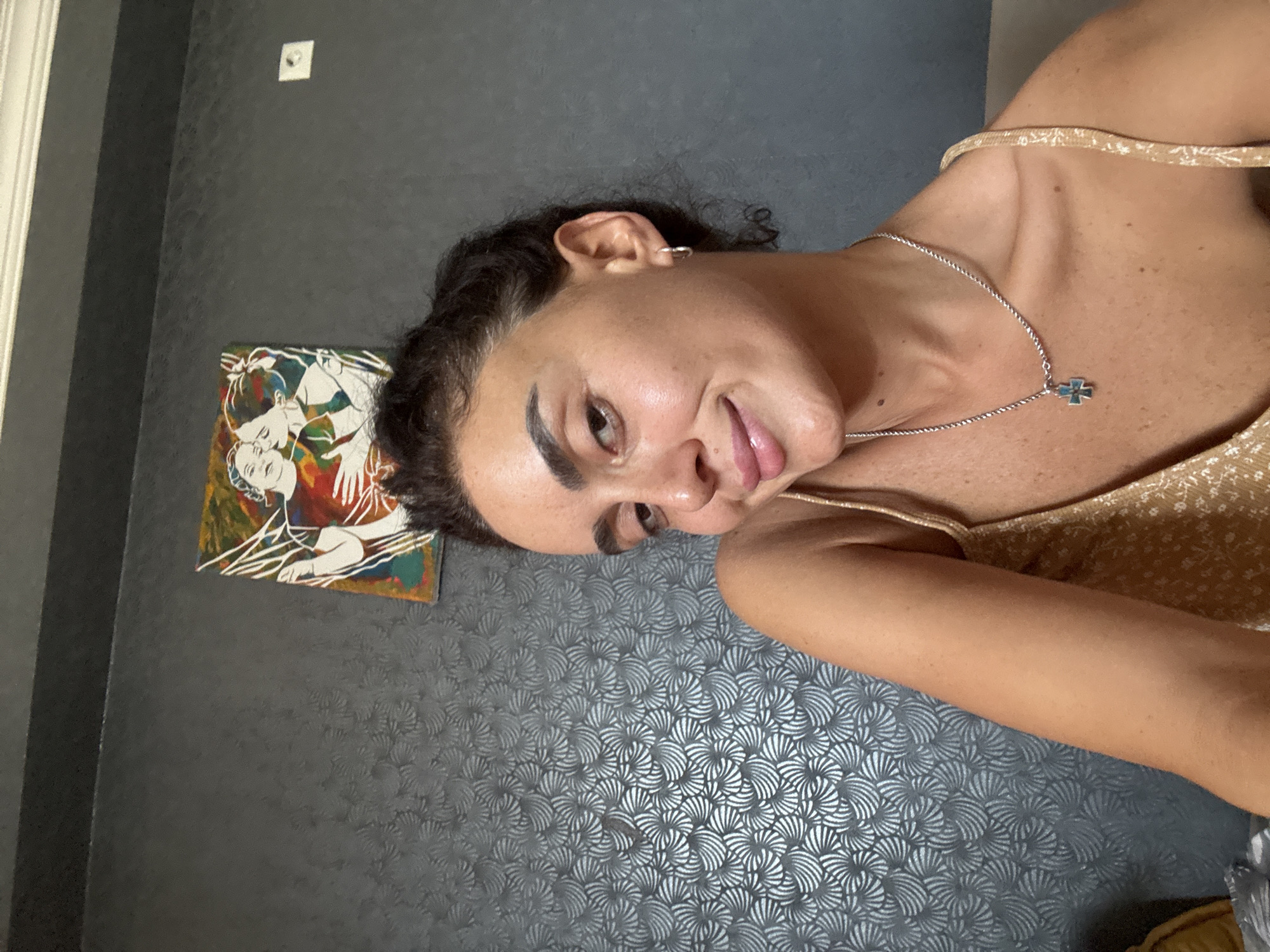 The image is a selfie taken indoors, likely in a residential setting in Tbilisi, Georgia. The subject is a woman with dark hair pulled back, looking towards the camera with a slight smile. She is wearing a gold-colored top with a delicate floral pattern and a silver necklace with a cross pendant. Her skin appears to be lightly tanned and dewy. Behind her, the wall has a textured gray wallpaper with a repeating pattern resembling stylized leaves or fans. On the wall above and to her left, there is a small, colorful abstract painting depicting two figures. The overall lighting suggests an indoor environment, possibly with natural light supplemented by artificial light. The scene conveys a casual and relaxed atmosphere.