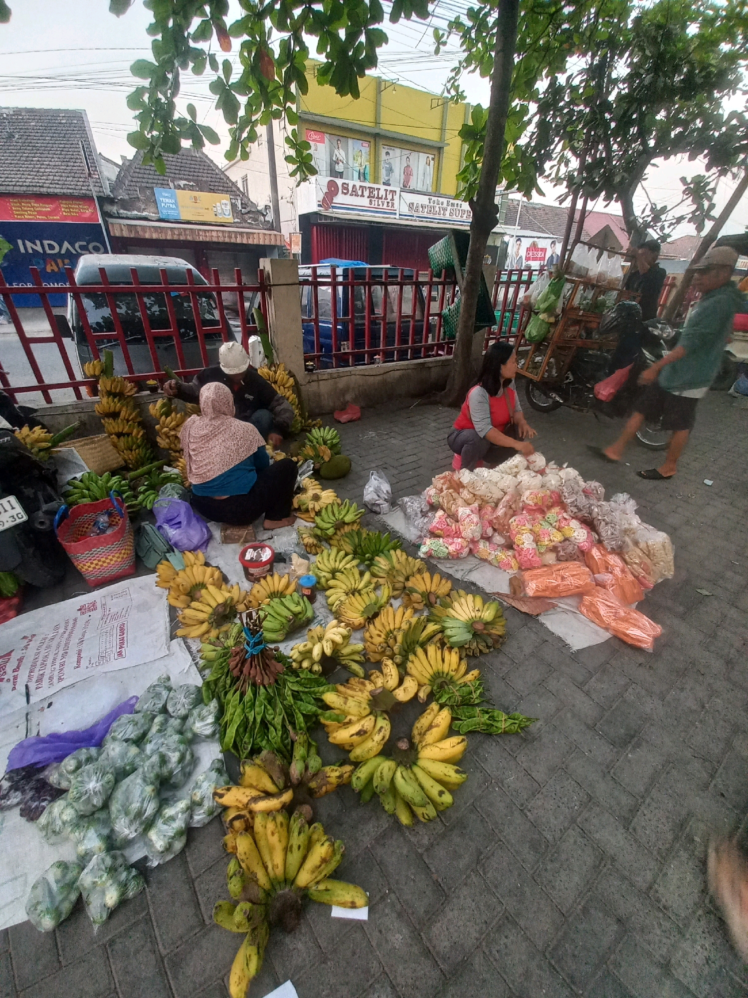 A street market scene in Baturetno, Indonesia, features a vendor selling fruits and snacks. Several bunches of bananas are displayed on the ground, some ripe yellow and others green. Adjacent to the bananas are bundles of green vegetables, likely a local variety. A separate pile consists of packaged snacks, some colorful and others in clear plastic. Two individuals are present; one, a woman in a red and gray shirt, is arranging the snacks, while another person, partially obscured, appears to be a customer. In the background, buildings with signs, including one for "INDACOR" and another that reads "SATELIT SUPER," line the street. A parked car and a motorcycle are also visible. The ground is paved with interlocking bricks, and the scene is partially shaded by leafy trees.