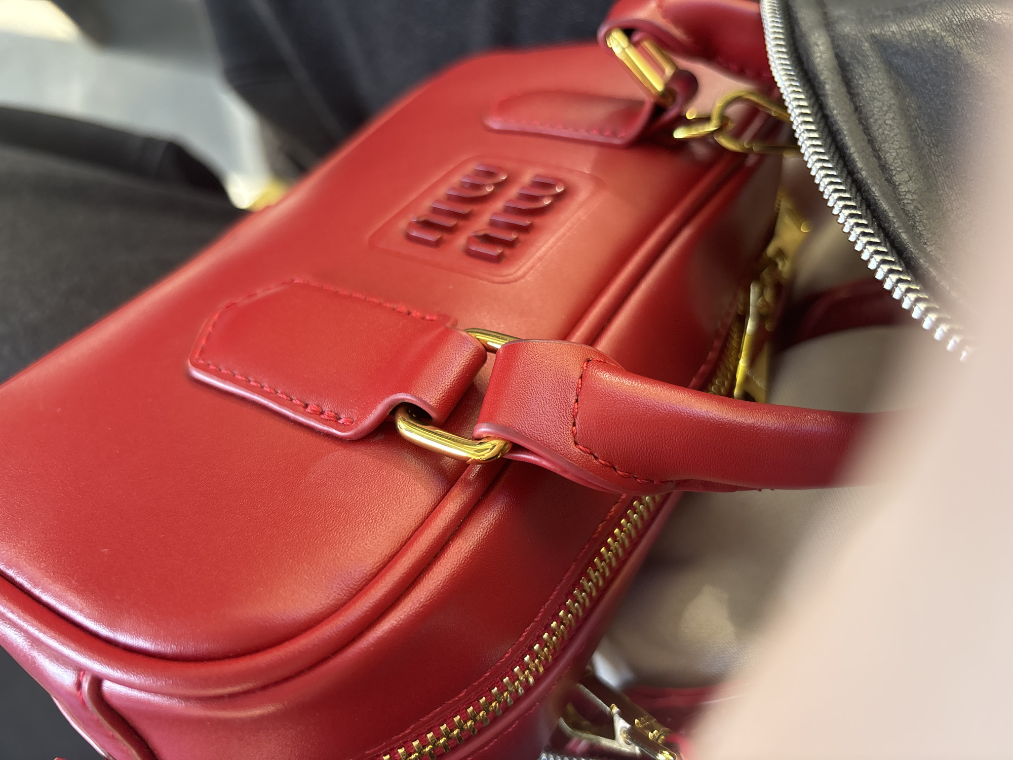 The image shows a close-up of a red leather handbag. The handbag has gold-colored hardware, including a zipper and attachments for a handle. The top of the bag features an embossed logo or design. Another black bag is partially visible on the right side of the image. The background appears blurry, suggesting the image was taken indoors. The location is not discernible from the image.