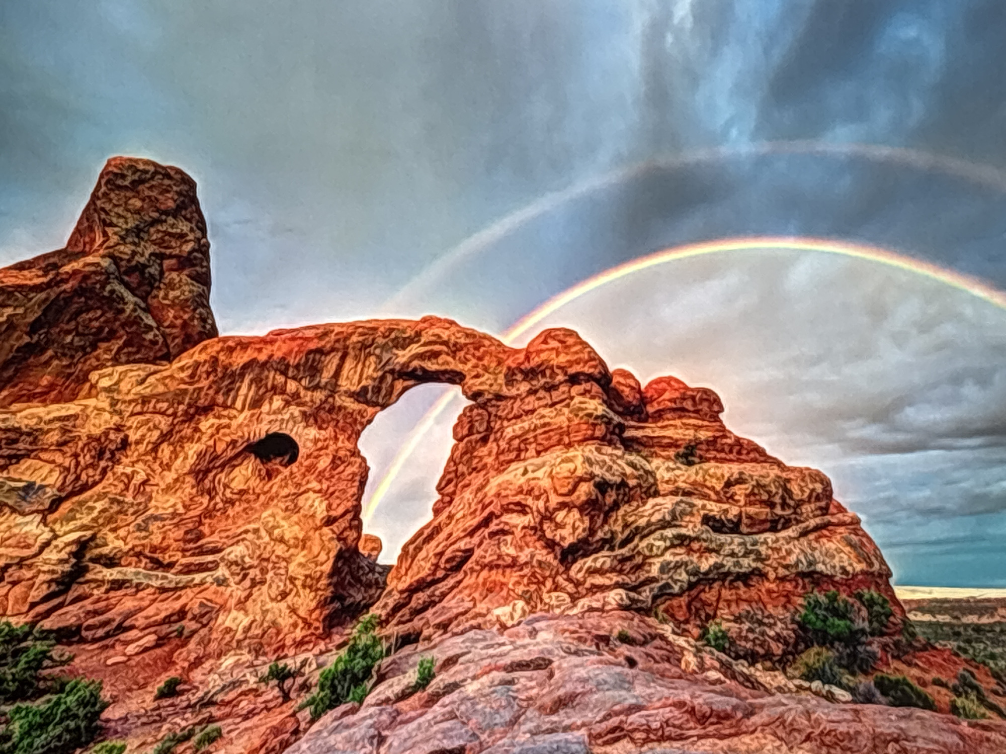 The image depicts a dramatic natural rock formation with an arch. The rock is reddish-orange and appears weathered, with layers and crevices visible. A dark opening is present in the left section of the rock formation. Green desert vegetation grows at the base of the rocks and on some of their surfaces.
Above the rock formation, a double rainbow is visible against a cloudy sky. The sky transitions from a lighter blue near the horizon to darker, heavier clouds overhead. The sunlight appears to be breaking through the clouds, illuminating the rainbow and the rock formation, suggesting a recent rain shower or the presence of mist.
The scene evokes a sense of awe and natural beauty. The ruggedness of the rocks contrasts with the ephemeral arc of the rainbows. The overall impression is one of a wild and majestic landscape.
There is no visible text in the image. The location context provided (Valladolid, Spain) does not appear to be supported by any specific visual cues within the image itself, which is more characteristic of desert landscapes found in regions like the American Southwest.