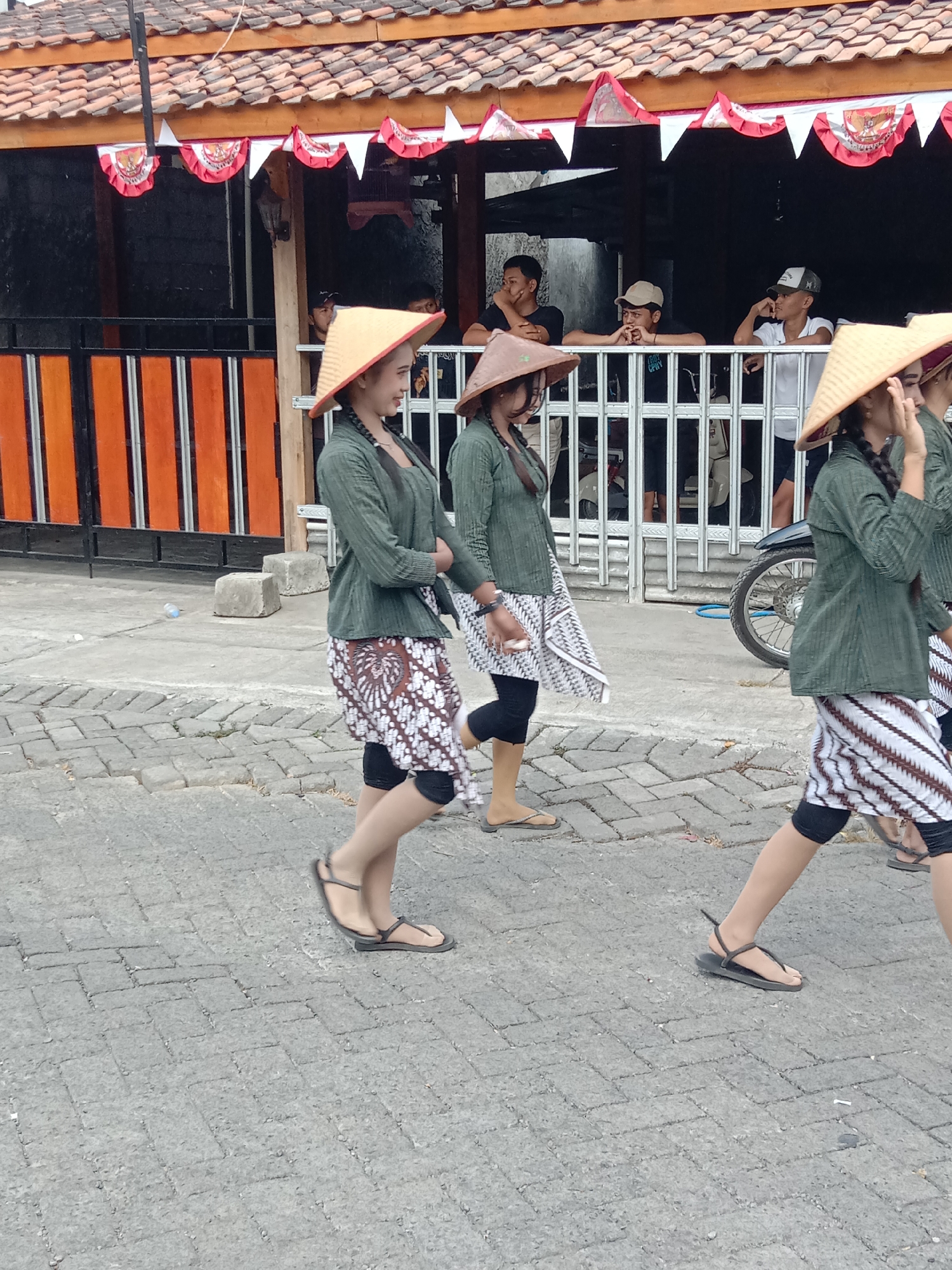 Three individuals, appearing to be women, are walking along a paved street in Wirosari, Indonesia. They are all wearing traditional conical hats and green tops with patterned skirts. The woman on the left has her hair in a braid and is wearing a batik skirt. The second woman, in the center, also has braided hair and is wearing a white skirt with brown patterns. The third woman, on the right, is also wearing a patterned skirt. Behind them, a building with a tiled roof and red and white decorative banners is visible. Several men are standing on a porch or balcony of the building, watching the procession. A motorcycle is parked to the right of the women. The ground is paved with cobblestones.
