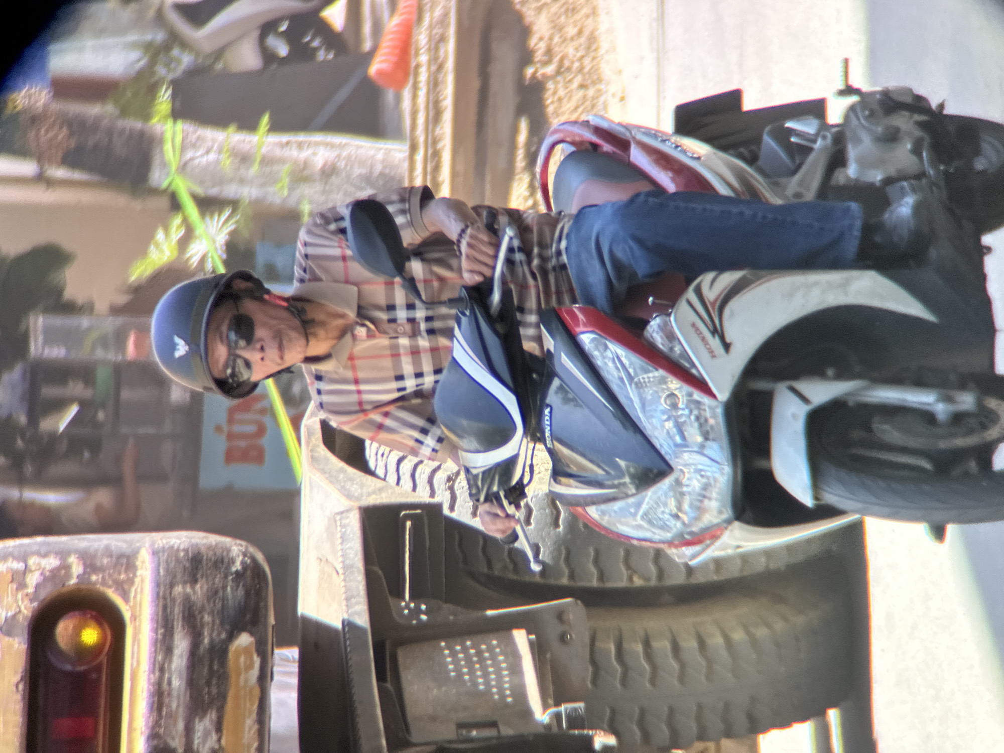 A man wearing a plaid shirt, sunglasses, and a helmet rides a scooter in Đà Nẵng, Vietnam. He is positioned in the foreground, with the side of a large truck to his left. The scooter is red and white, with the word "HONDA" visible on its side. The man's right hand is on the handlebar. The background shows some blurred foliage and a sign with Vietnamese text that reads "BÚN". The scene appears to be a street.