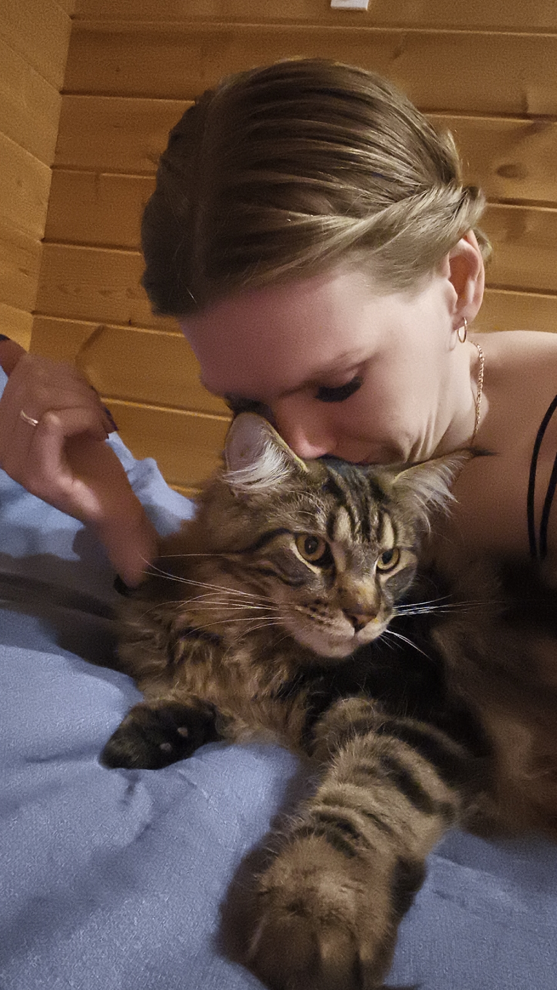 A fair-skinned woman with brown hair pulled back in a twisted updo is pictured from the shoulders up in close proximity to a brown tabby cat lying on a blue blanket or bedspread. She is kissing the cat on the head and scratching the cat's chest with her left hand, which is adorned with a ring on the ring finger. The woman is wearing a thin, dark-colored tank top and jewelry, including hoop earrings and a delicate necklace. The cat has amber eyes and prominent whiskers. Behind the woman is a wall constructed of horizontal wooden planks, possibly indicating an interior wood-paneled wall. The location is identified as Русский Вожой, Russia, although there are no visible landmarks to confirm this. The scene portrays affection between a woman and her pet cat.