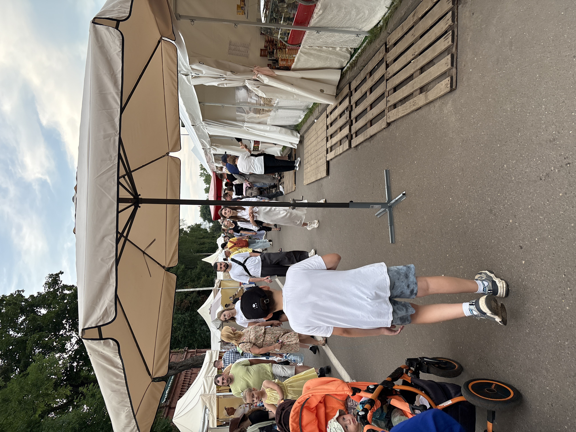 The image depicts an outdoor market scene, likely in the city of Vitsebsk, Belarus, given the context provided. A large, beige umbrella with a black trim dominates the foreground, casting a shadow over a paved walkway. In the center of the frame, a young boy wearing a white t-shirt and patterned shorts walks away from the viewer, his back to the camera. To the left, an orange stroller is visible with a young child inside wearing a hat. Further into the background, a diverse crowd of people mill about between white market tents, suggesting a lively event or fair. Several tents are set up along the right side of the walkway, displaying goods on shelves and tables. The ground is paved with asphalt, and wooden pallets are laid out in front of some of the tents. Trees and a building with a brick facade are visible in the background, hinting at the surrounding urban environment. The sky above is partly cloudy.