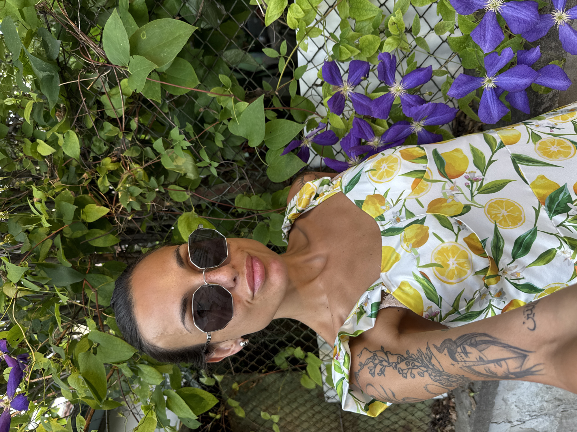 In Pleven, Bulgaria, a woman is pictured outside. She is wearing a white dress with a lemon print and sunglasses. She has dark hair pulled back and a tattoo on her left arm. She is standing in front of a green vine-covered fence and purple flowers. The lighting suggests it is daytime.