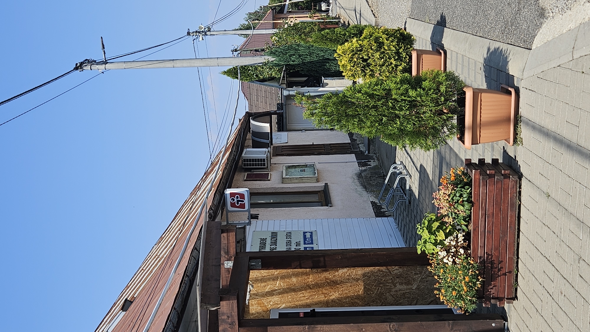 The image shows a street scene in Vyšné Nemecké, Slovakia. In the foreground, there is a wooden planter box filled with orange and green flowers. To the right of the planter box, there are two potted evergreen trees. The trees are in brown plastic pots, and the one on the right is larger and bushier than the one on the left.
Behind the planters and trees, there is a row of buildings. The building in the foreground has a light-colored facade and a rusty metal roof. There is a sign on the building with a red and white logo. To the right of this building, there are more buildings with tiled roofs, some of which are brown. There are also power lines and a lamppost visible in the background. The street is paved with cobblestones. The scene appears to be a commercial area, possibly with shops or offices. The sky is clear and blue.