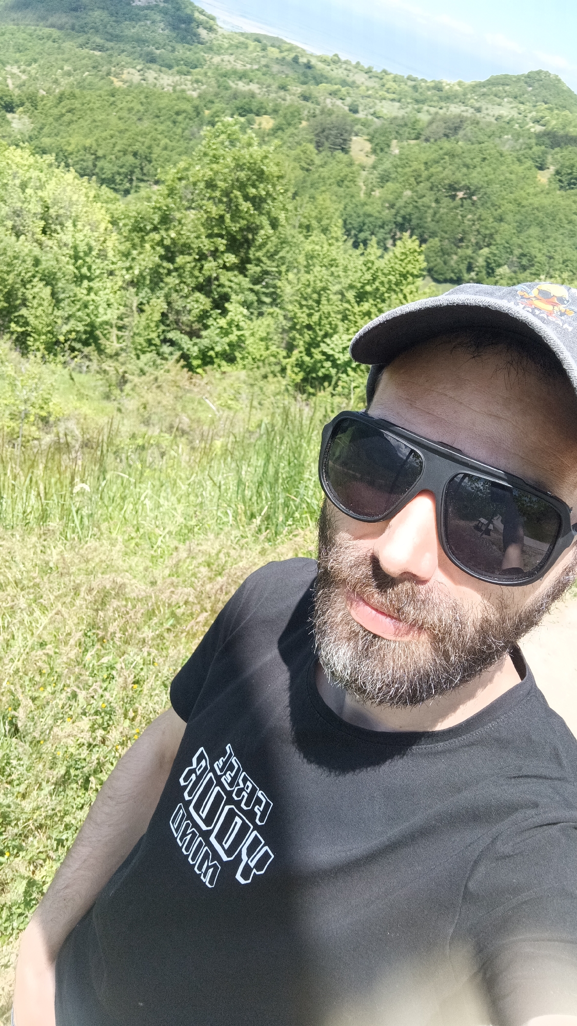 A selfie shows a man with a beard, wearing sunglasses and a baseball cap, outdoors. He is wearing a black t-shirt with a white logo. The background features lush green trees and vegetation. In the distance, a mountain or hill is visible under a blue sky. The location is Oreshets, Bulgaria.
