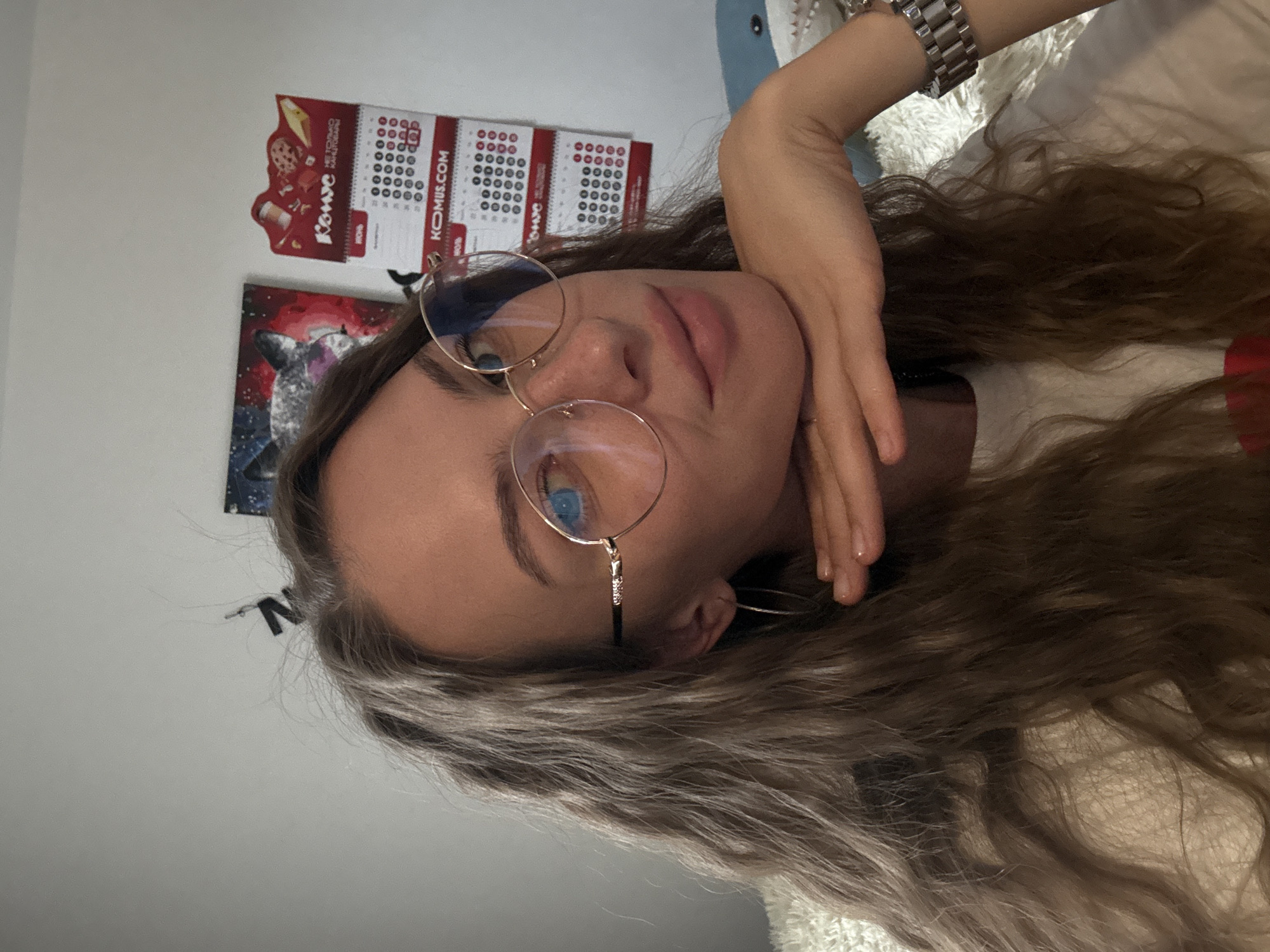 The image is a close-up, selfie-style photograph of a young woman with long, wavy brown hair and light-colored eyes wearing round, gold-rimmed glasses. She is resting her chin on her left hand, which is positioned in front of her mouth. Her right arm, adorned with a silver wristwatch, is bent upwards. She is wearing a white t-shirt. In the background, a white wall is visible, with a framed artwork depicting a wolf on the left and a wall calendar featuring images of food items and dates on the right. The calendar appears to be from "KOMUS.COM". A plush white blanket or rug is visible on the lower left of the frame, and a portion of a blue shark-shaped object is visible on the right. The setting appears to be an indoor residential space, possibly a bedroom. No specific location in the city of Perm, Russia, is identifiable from the image.