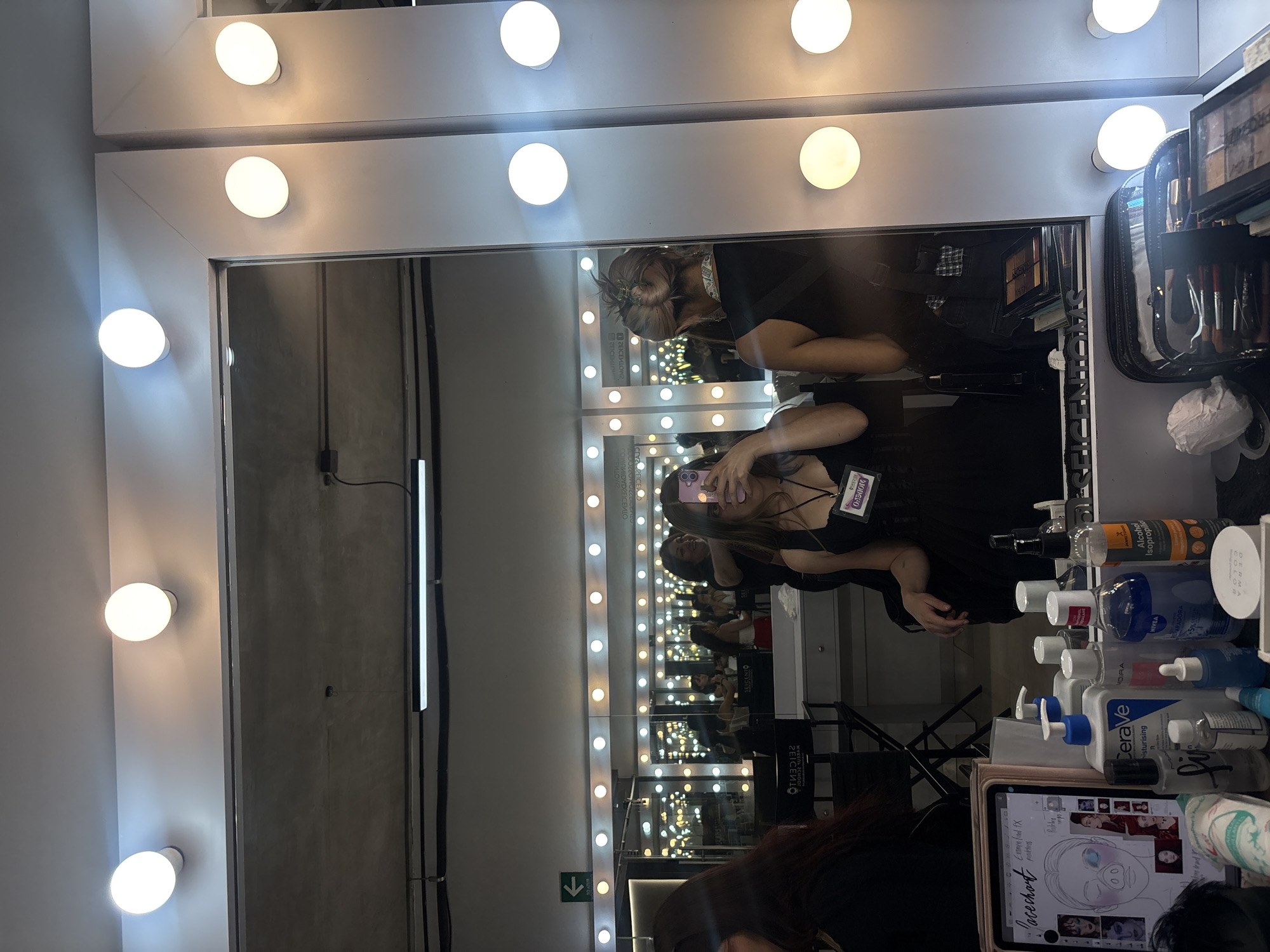 The image captures a makeup station, likely in a professional setting, as suggested by the multiple illuminated makeup mirrors reflecting rows of similar stations. There are at least two people visible in the mirrors: one holding a cell phone taking the photo, and another in the background working on the makeup. The makeup station has an assortment of skincare products and makeup supplies, including Cerave lotion, Nivea cream, and alcohol. A tablet showing a face chart is also present. The lighting indicates a bright environment intended for makeup application. The word "SEICENTO" is mirrored and readable. The presence of an exit sign and the general layout suggest a location in Mexico City, Mexico.