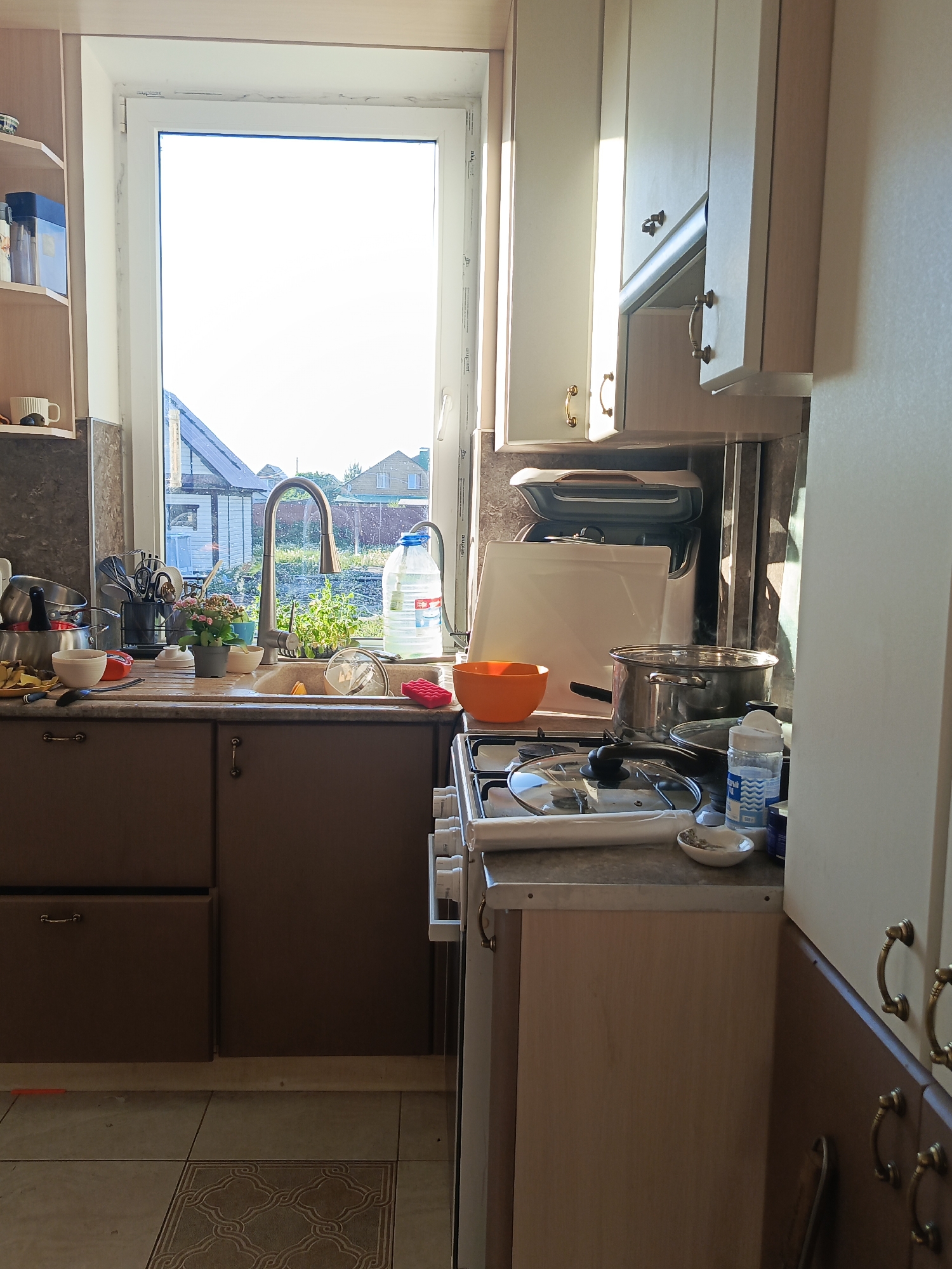 This image depicts a cluttered kitchen in Unknown, Unknown. The scene centers around a sink filled with dishes, pots, pans, and various kitchen utensils. Adjacent to the sink is a stove top with additional cooking items and a partially visible orange bowl. A large window behind the sink offers a view of a residential area. The cabinets above the sink and to the right are light colored wood. The floor is tiled in a patterned design. The overall scene suggests a space actively in use.