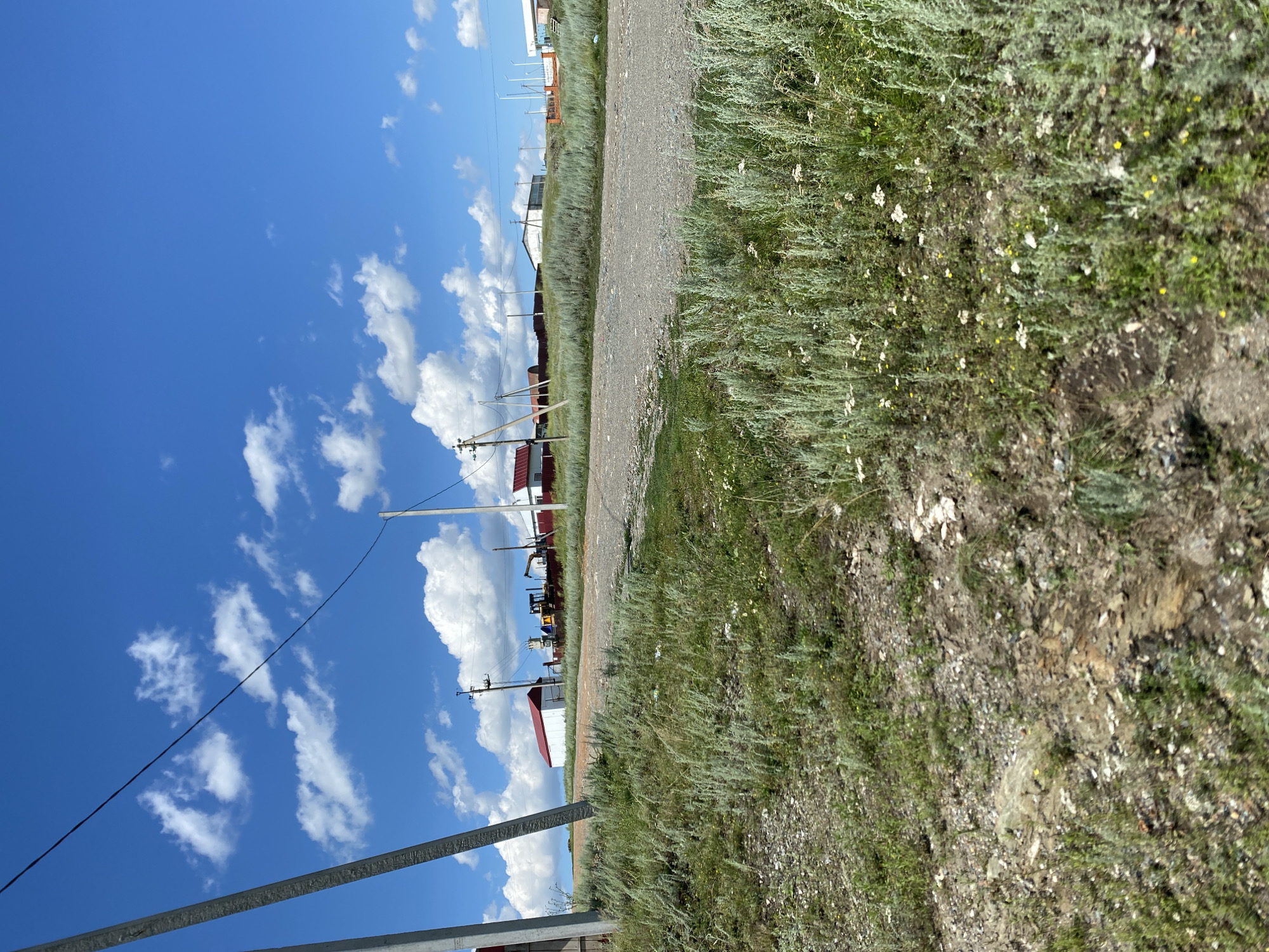 The image depicts a rural scene with a dirt road leading towards a collection of buildings. The sky is bright blue with scattered white clouds. The road is flanked by tall, green grass and wildflowers. In the distance, several buildings with red roofs are visible, along with what appears to be construction equipment, including a yellow excavator. Power lines and utility poles are also present in the scene. The overall impression is one of a sunny day in a semi-arid, rural environment, likely in or around Yuldybayevo, Russia, given the provided context. There are no people visible in the image.