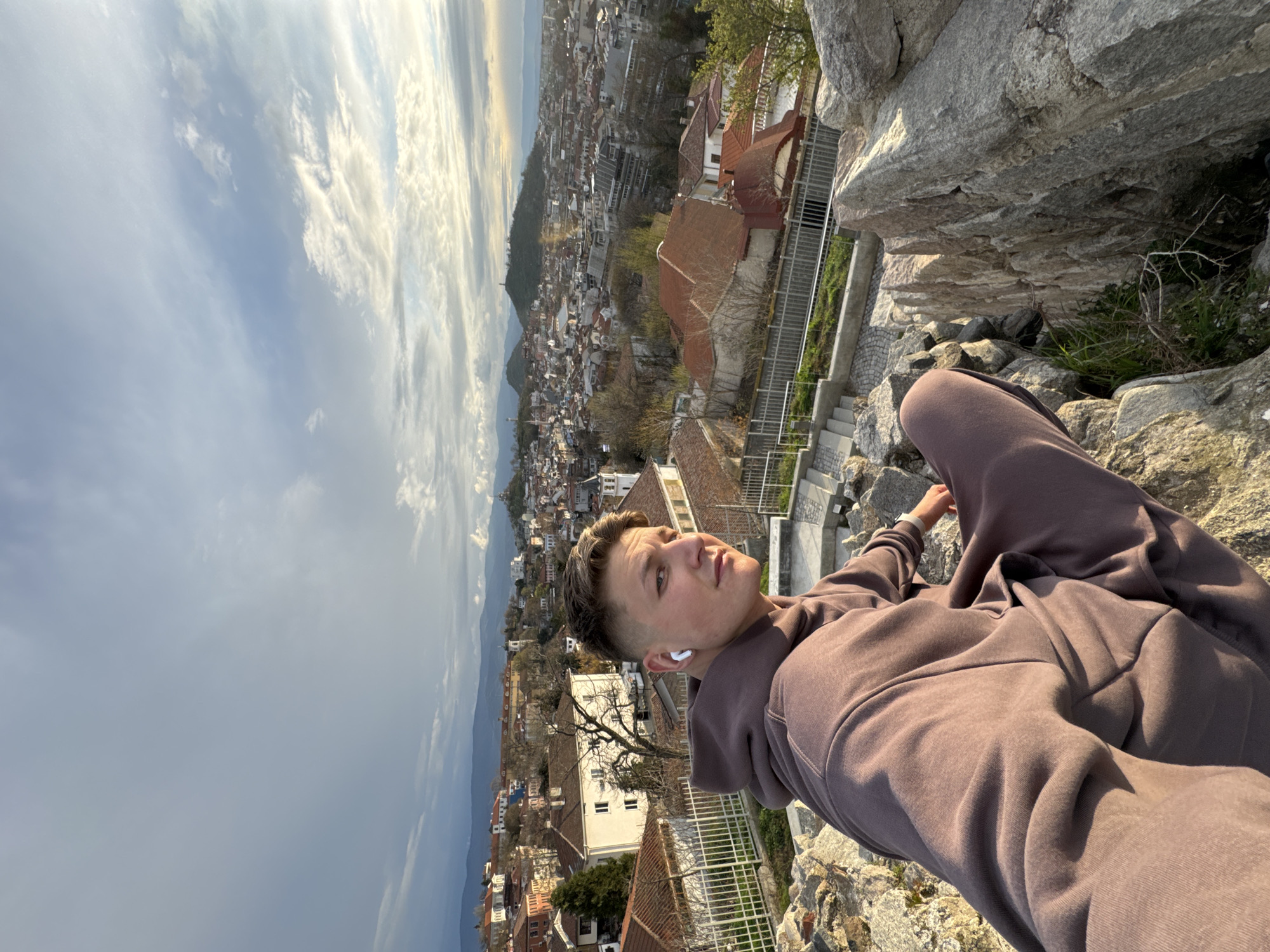 A medium-shot portrait taken from a high angle depicts a light-skinned young man with short, styled hair, wearing a brown hoodie and sweatpants, sitting cross-legged on a rocky outcrop. He has a wireless earbud in his left ear and is gazing upwards and to the right.
In the background is a cityscape, likely Plovdiv, Bulgaria. Visible are buildings with red-tiled roofs, along with a small mountain in the distance. The sky is visible in the upper left corner, with a mix of sunlight and clouds.
To the right of the figure are gray stone rocks and a set of stairs. The photograph has been rotated counter-clockwise, so the perspective is tilted. The location appears to be Nebet Tepe hill in Plovdiv, Bulgaria, based on the vantage point and cityscape visible in the background.