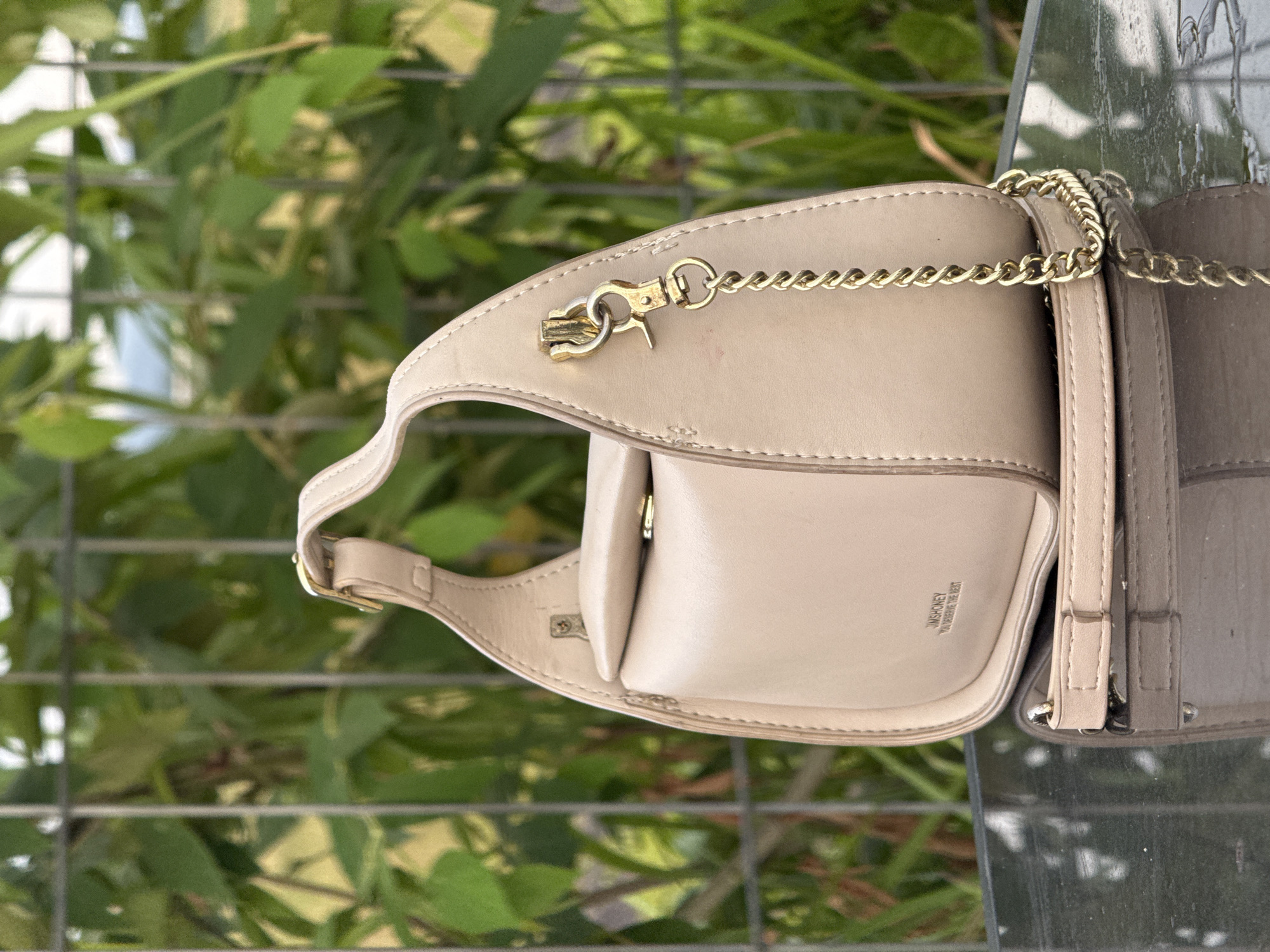 A light beige handbag with a gold chain strap is displayed on a reflective dark surface. The handbag has a structured shape with a flap closure and subtle stitching. A gold clasp and chain are attached to the side of the bag. The background consists of green foliage behind a metal fence. The scene appears to be outdoors, possibly in a garden or park setting. There is no indication of the city of Cotmalem, Indonesia within the image.
