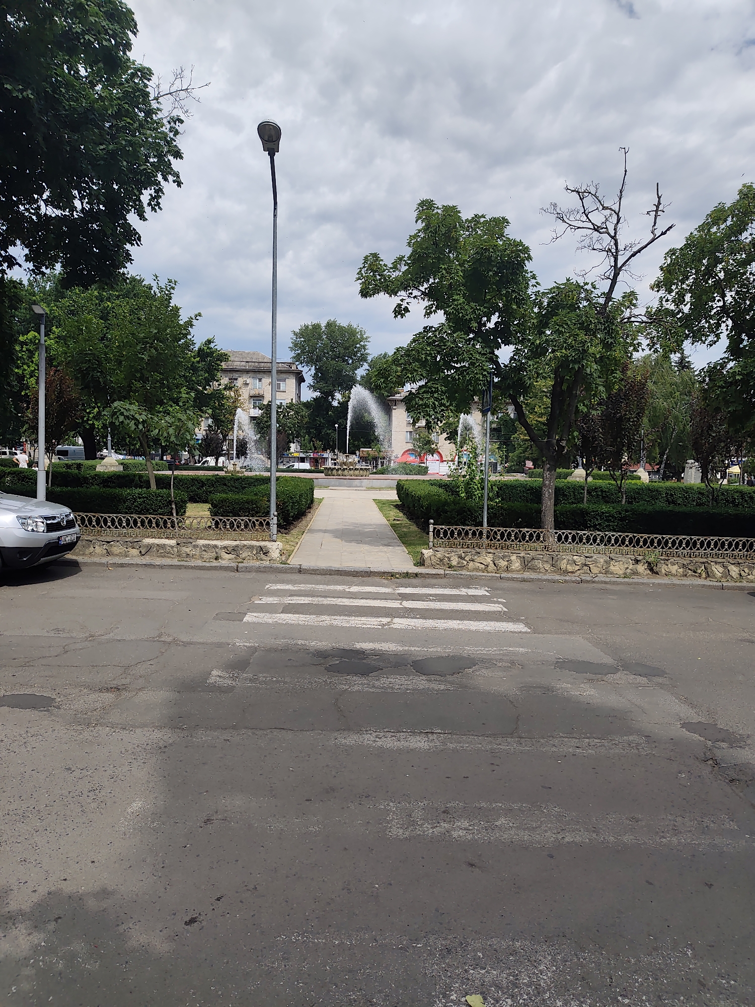 This image depicts a street scene in Chișinău, Moldova. In the foreground, a crosswalk with white stripes is visible on a cracked asphalt road. To the left of the crosswalk, a silver car is parked. Beyond the road, a paved walkway leads into a park area. The park features manicured hedges, several trees, and fountains spraying water. In the background, a building with multiple windows and balconies can be seen, as well as streetlights and a cloudy sky. The overall environment suggests an urban public space.