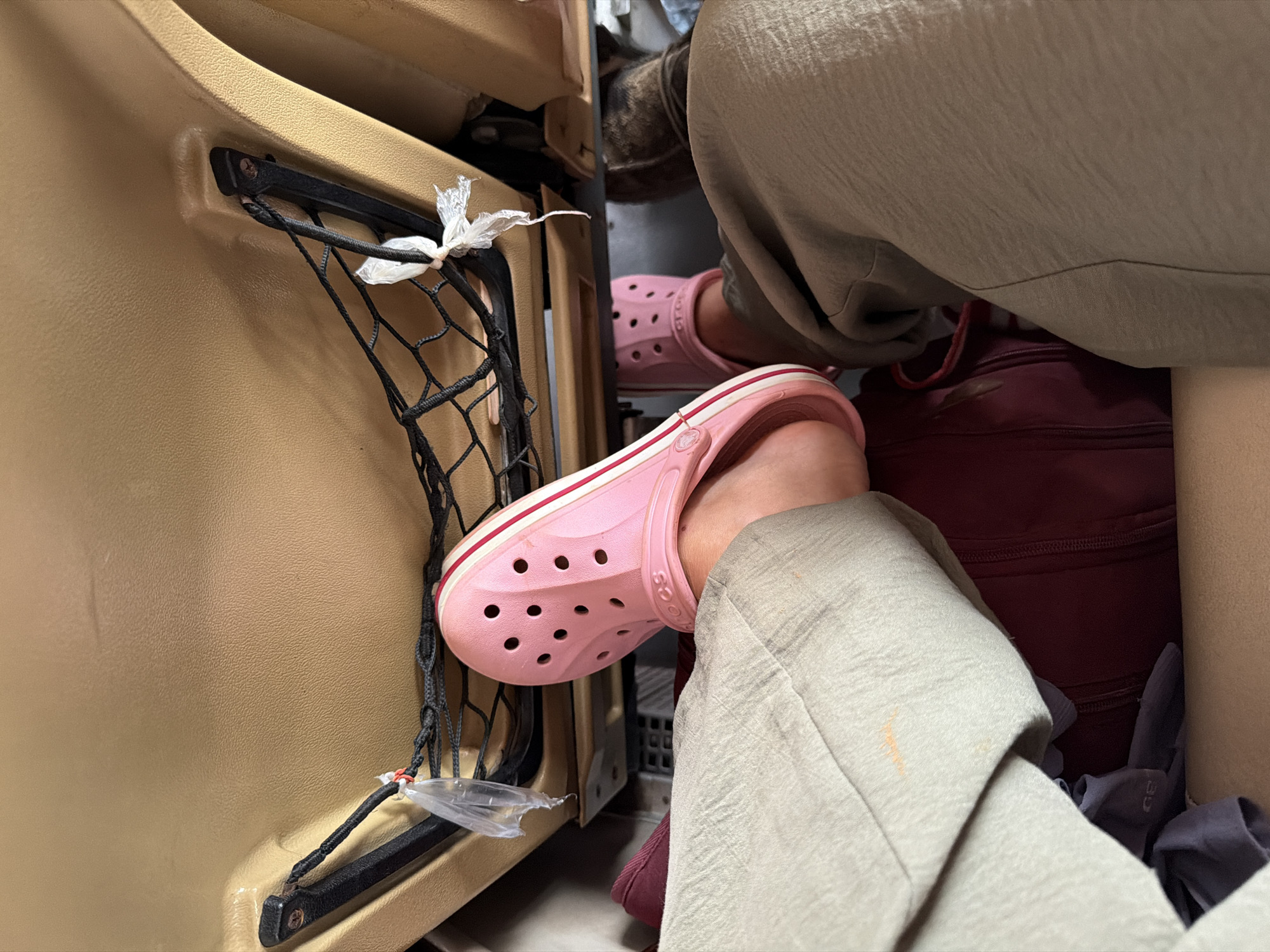 The image shows a close-up, first-person perspective of a person's legs and feet, wearing pink Crocs and light-colored pants, resting in a vehicle's passenger seat. The backdrop reveals the interior of a car, specifically the back of a front seat with a mesh pocket. A maroon bag is visible beneath the person's legs. The environment suggests a travel or commute context, likely within the city of Phakhao, Laos, given the inferred location.