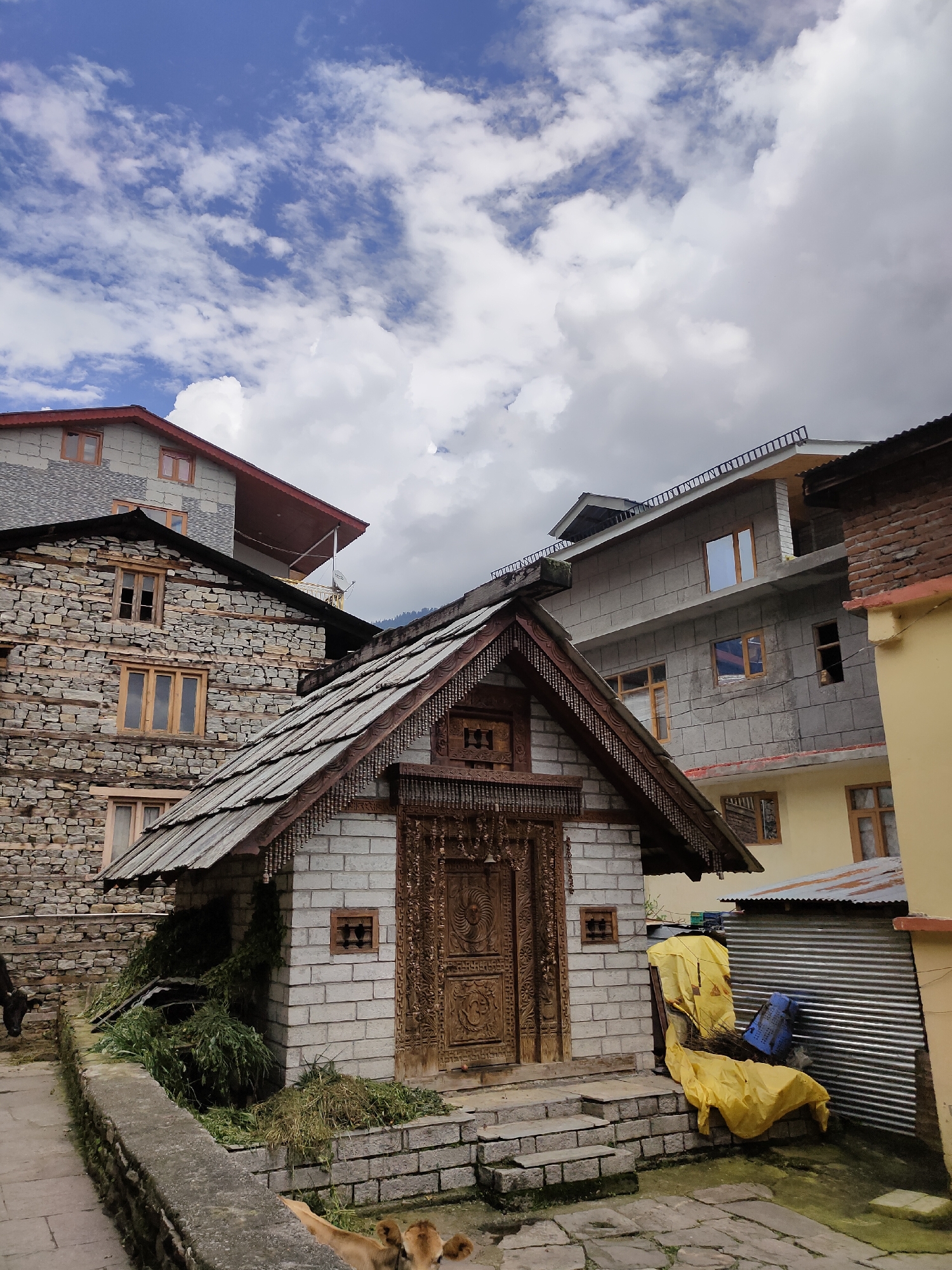 In Siyal, India, a collection of multi-story buildings with varied architectural styles and materials dominates the scene. The foreground features a small, ornate structure with a wooden door and roof, constructed from light-colored bricks. Adjacent to this, a corrugated metal shed is covered with a bright yellow tarp, and a blue plastic crate rests against it. To the left of the small structure, a stone wall runs along a paved walkway, with a cow visible in the lower right and another to the left in the distance. Behind the small structure and stone wall, a larger building constructed of rough-hewn stones with wooden window frames is visible. Further in the background, more modern buildings with concrete facades and windows are present, interspersed with older structures. The sky above is a mixture of blue and white clouds.