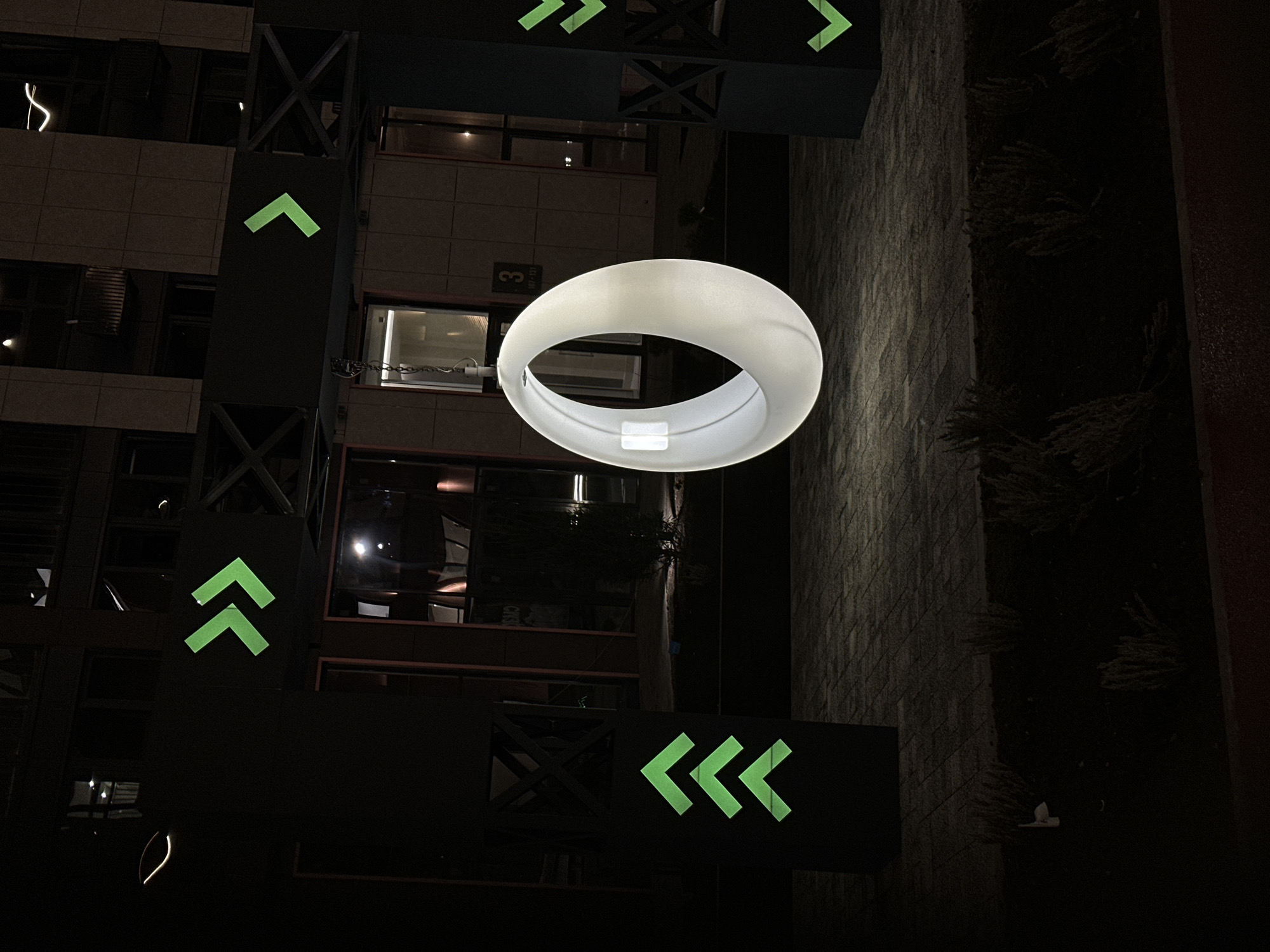 The image depicts a nighttime scene in what appears to be Rostov-on-Don, Russia. Tall buildings surround an open area. The buildings feature architectural elements like external metal supports and various window configurations.
A prominent illuminated ring-shaped fixture hangs in the center of the open space. The scene includes green illuminated directional signs that have arrows that are point upwards and leftwards.
A textured vertical wall, possibly made of stone or brick, runs vertically on the right side of the frame. Plant life is interspersed along the wall. The photograph is taken from a slightly downward angle, possibly from a high vantage point. The overall ambiance is dark and somewhat surreal.