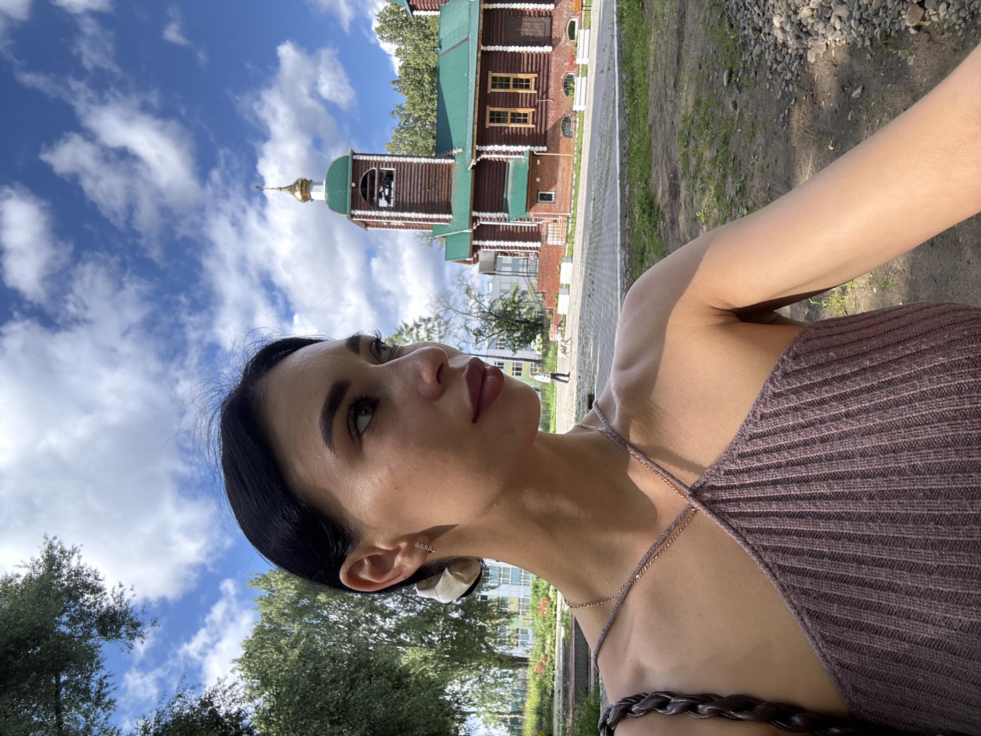 The media file features a young woman in the foreground, looking to her left and upwards. She has dark hair and is wearing a ribbed, sleeveless top with thin straps, and a delicate necklace. Her earlobe has a small, delicate earring. The background is a scene with a traditional wooden church building with a green roof and a golden dome, to the left of the image. To the right of the church is a paved walkway and a grassy area with some gravel. Trees and a blue sky with white clouds complete the outdoor setting. The lighting suggests it is daytime, possibly late afternoon given the shadows. The overall mood seems relaxed and observational. No text is visible.