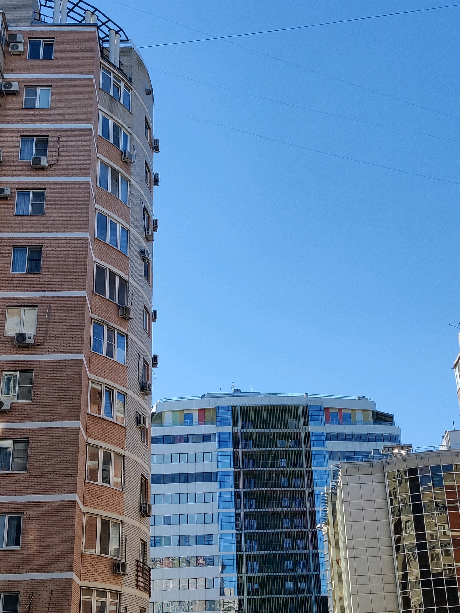 The image shows a vertical shot of several modern buildings against a clear blue sky. On the left, a curved apartment building made of reddish-brown brick is visible, with white horizontal bands and many windows, each with an air conditioning unit mounted below it. To the right of the brick building, a taller, white building with many windows and balconies can be seen. Further to the right and behind the other two, a glass-fronted building with a colorful, undulating roofline is partially visible. The overall scene depicts an urban environment on a bright, sunny day.
