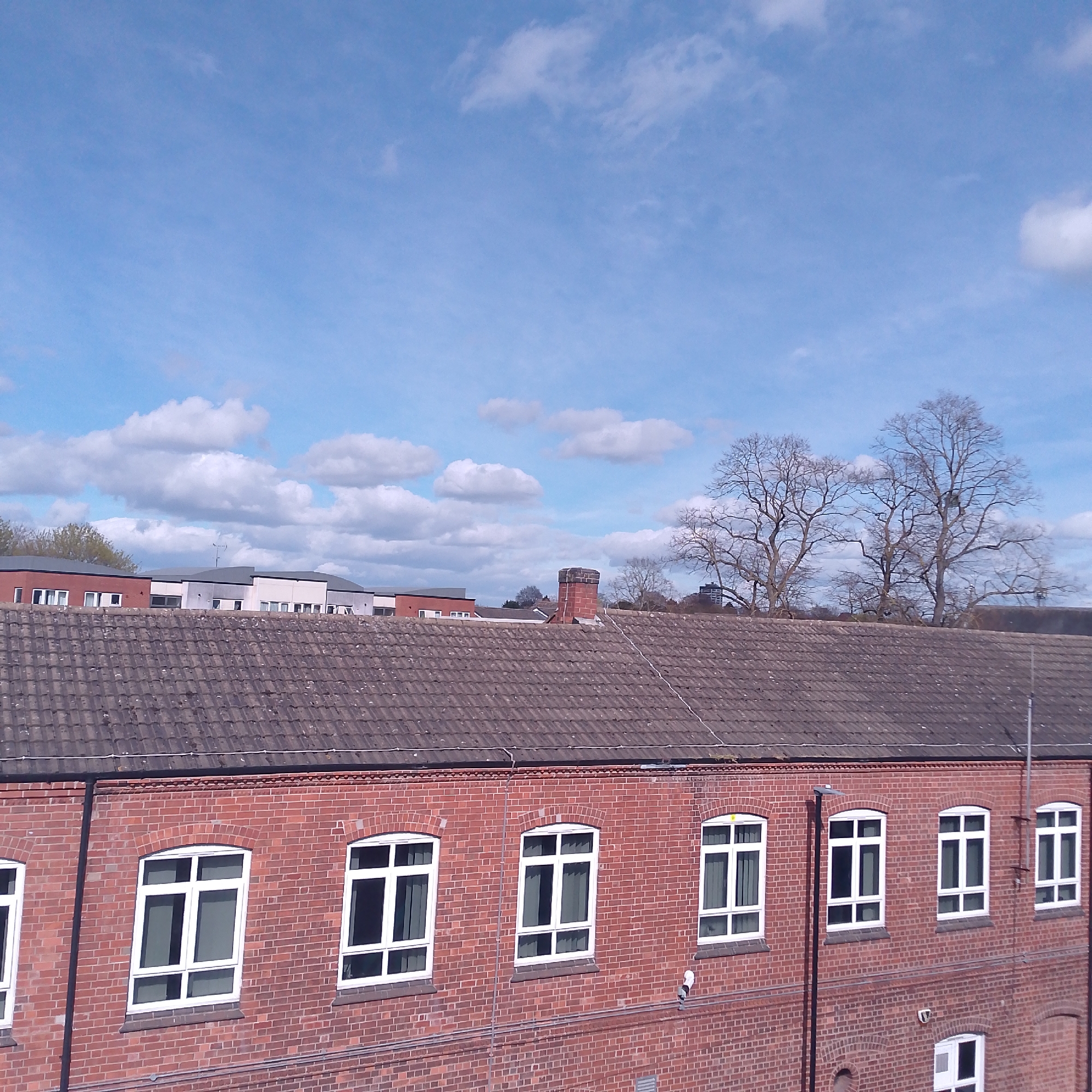 The image depicts a red brick building with a dark tiled roof, located in Coventry, United Kingdom. The building has several white-framed windows, symmetrically arranged. The roof also has a brick chimney. Behind the building are trees without leaves and other buildings with various roof colors. The sky is blue with scattered white clouds. There are no people visible in the image.