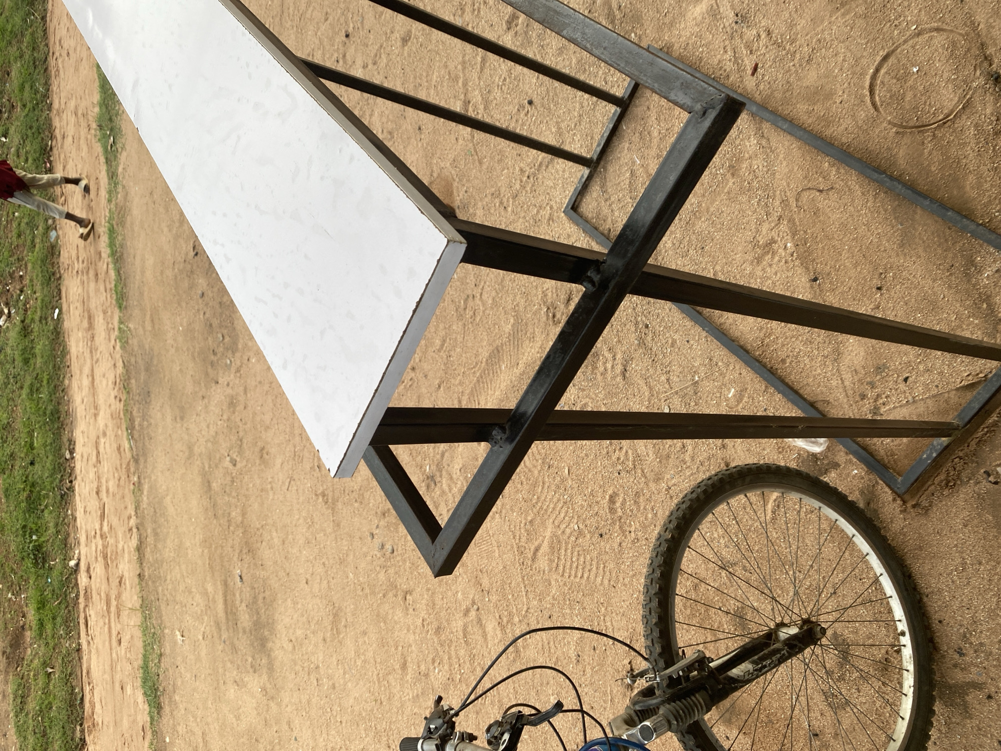 A bicycle wheel and part of its frame are visible on the left side of the frame, positioned on sandy ground. To the right, a black metal rack with a white surface extends diagonally across the image. In the background, a person wearing a red top and light-colored pants walks on a dirt path lined with grass. The scene appears to be outdoors, likely in Jalingo, Nigeria, given the sandy terrain and the presence of a person in a casual setting.