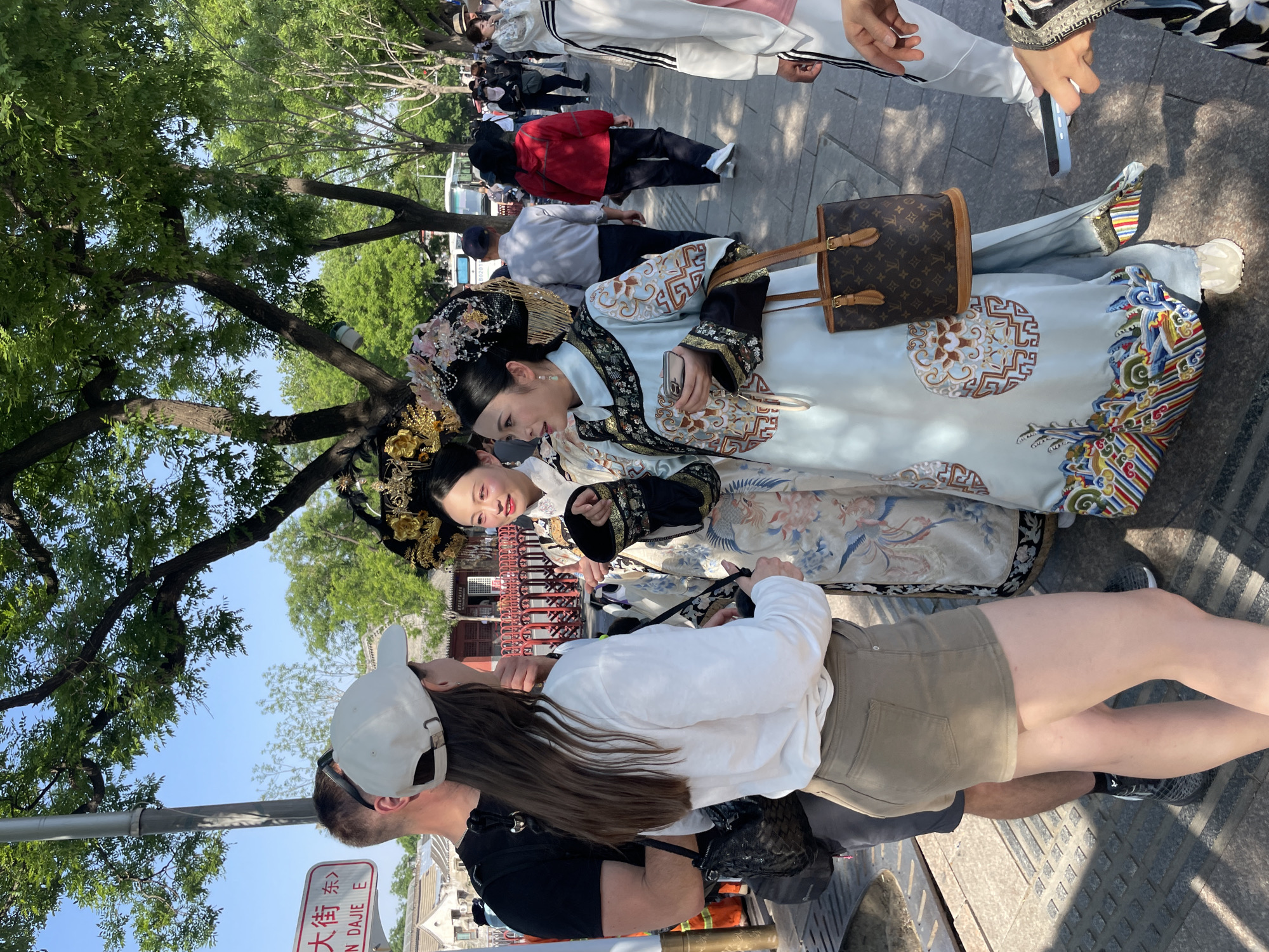 The image captures a scene in the Dongcheng District of Beijing, China. In the foreground, a couple is standing on a paved walkway near a sign with Chinese characters that include “DAJIE” which translates to “Big Street”. The man is wearing a black shirt and a white baseball cap. The woman is wearing a white long sleeve shirt and khaki shorts. They appear to be interacting with two women who are dressed in traditional Chinese clothing, including elaborate headdresses. One of the women is holding a smartphone. An LV (Louis Vuitton) purse is present. In the background, other people can be seen walking along the walkway. A tree is visible on the left.