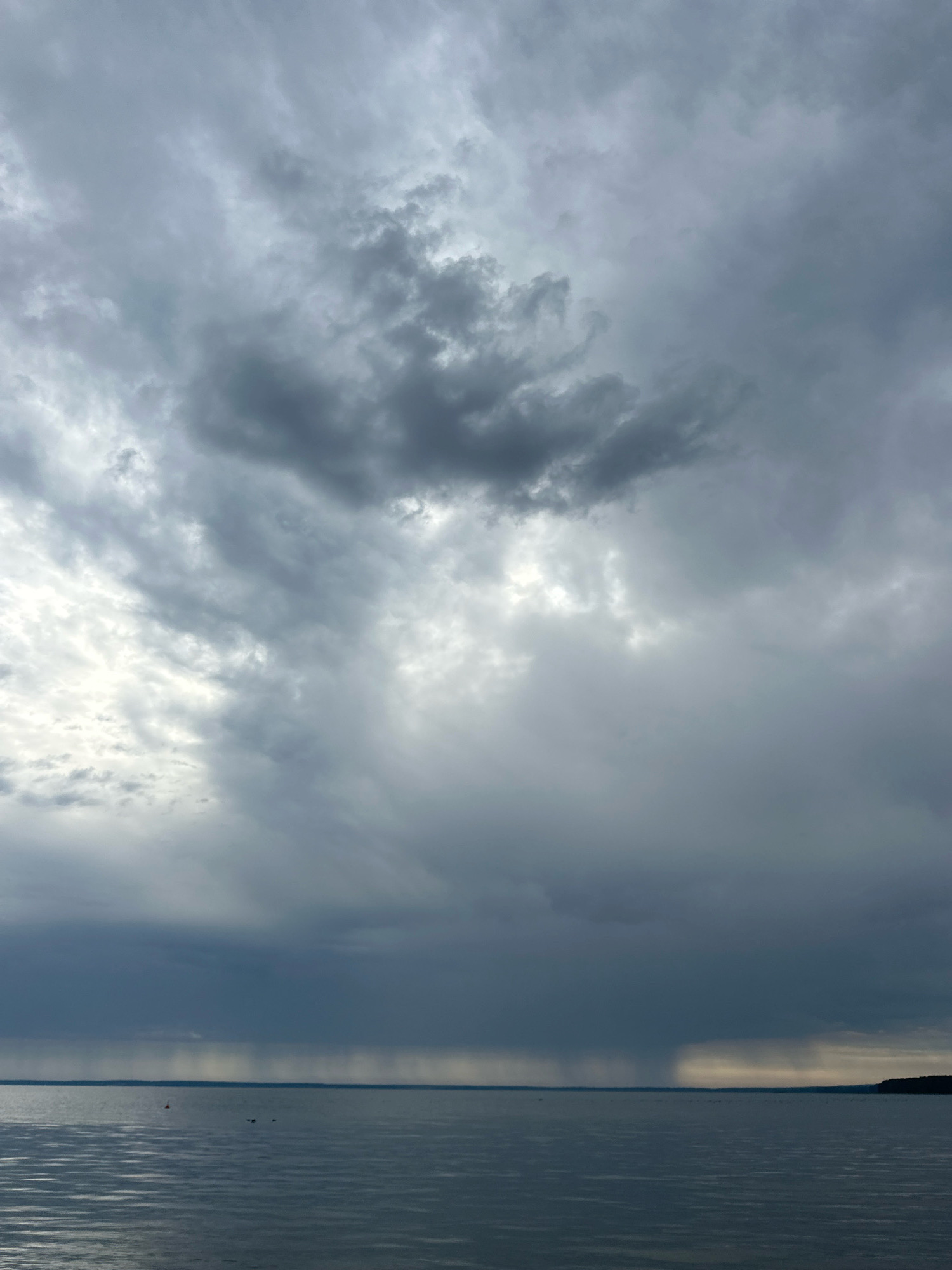 The image depicts a wide, calm body of water under a dramatic, overcast sky. Heavy, dark clouds dominate the upper portion of the frame, with lighter patches of sky visible through breaks in the cloud cover. In the distance, a horizontal line of rain is visible falling over the water, creating a blurred effect against the horizon. The water's surface is relatively smooth, with subtle ripples reflecting the muted light. The far edge of the water is bordered by a dark, tree-lined shoreline on the right side of the image. The scene evokes a sense of impending weather and the vastness of the natural environment. The location is identified as Нарацкі сельскі Савет, Belarus. There are no people or specific objects clearly discernible in the foreground or midground of the image.