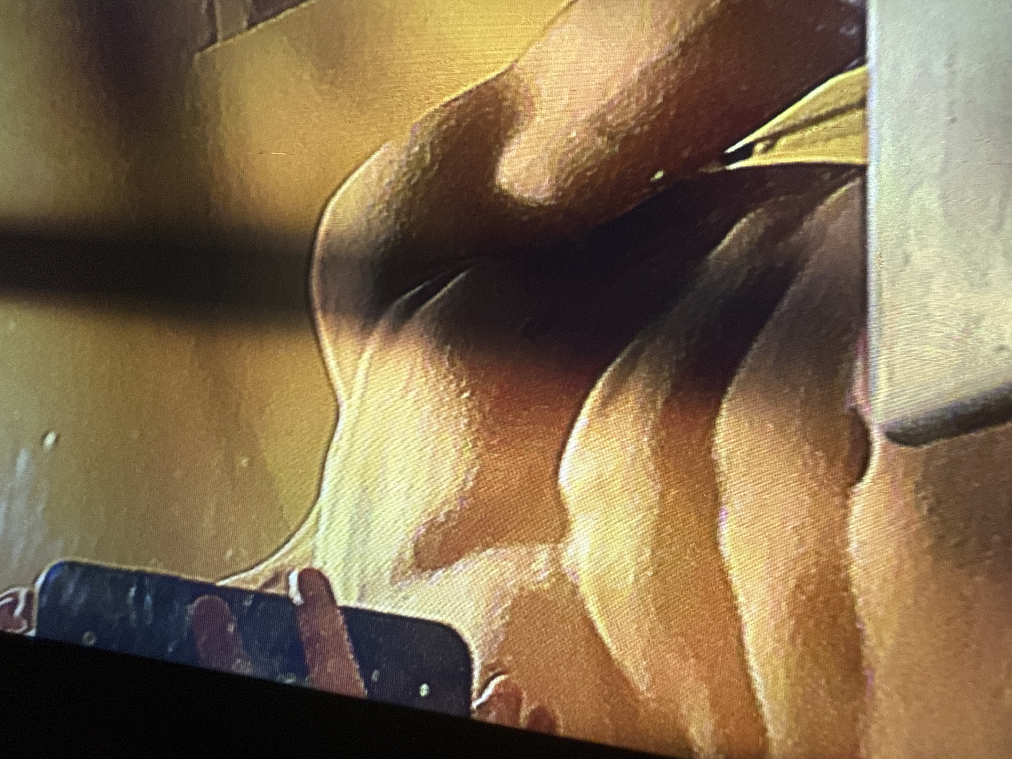 The image is a close-up, low-angle shot of a person's back and buttocks, illuminated by warm, golden light. The person appears to be wearing a light-colored bikini bottom. The focus is on the muscular definition of the back and the curve of the buttocks. The background is a blurry wash of golden-brown and dark shadows. No recognizable city landmarks or specific locations in Sofia, Bulgaria, can be identified.