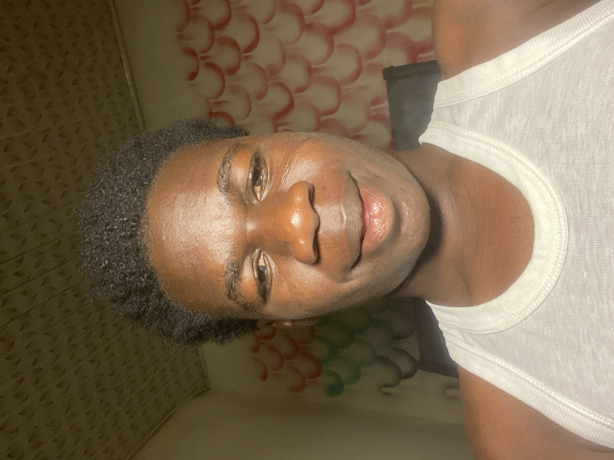 A close-up, portrait-style image of a Black person with short, dark hair wearing a white tank top. The person appears to be indoors, based on the patterned wallpaper behind them. The wallpaper features a design of red and white overlapping shapes that resemble scales or petals. There's also a section of wall with a different pattern, possibly mosaic tiles, in the lower-left corner. The location is Mararaba, Nigeria is not discernible from the image.
