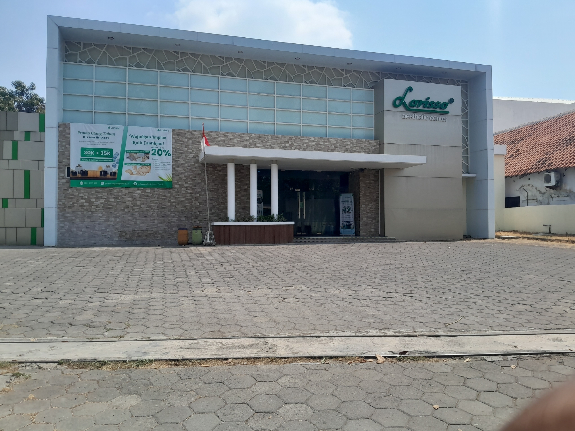 The image shows the front exterior of a modern building, identified as "Larissa Aesthetic Center" in Tegal, Indonesia. The building features a facade with large glass panels on the upper level, adorned with a geometric pattern. The lower portion of the building is covered in textured stone tiles. A prominent green sign with the "Larissa" logo is visible on the right side of the building.
In front of the building, a paved area with hexagonal concrete blocks serves as a walkway and parking space. A large banner is displayed on the left side of the building, promoting "Promo Ulang Tahun It's Your Birthday" and "Wujudkan Impian Kulit Cantikmu!" with a 20% discount offer. The banner also includes images of cosmetic products and a baby.
Two cylindrical trash bins, one orange and one green, are situated to the left of the building's entrance. The entrance itself consists of glass doors. The overall environment appears to be a bright, sunny day with a clear blue sky. To the right of the aesthetic center, a building with a terracotta tiled roof is partially visible, suggesting a mix of architectural styles in the area. No people are clearly visible in the immediate foreground or around the building's entrance.