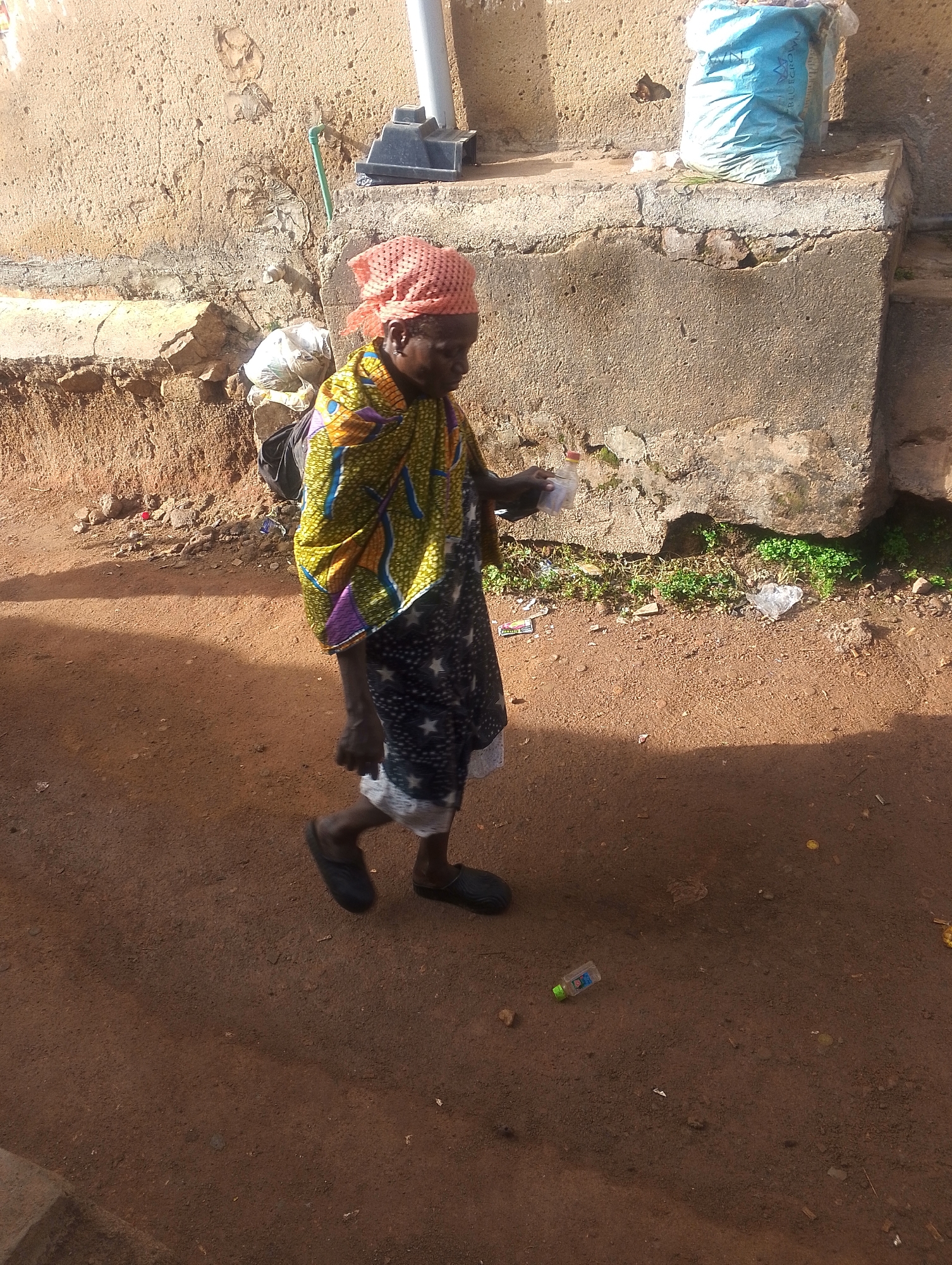An elderly woman wearing a headscarf, a colorful patterned shawl, and a dark dress with white stars walks on a dirt road. She carries a bundle on her back and holds something small and white in her right hand. The road is littered with trash, including a small plastic bottle. Behind her is a concrete wall with a water pipe. A large blue bag sits on the wall. The location appears to be in a residential area of Jos, Nigeria, given the style of construction and the state of the unpaved road.