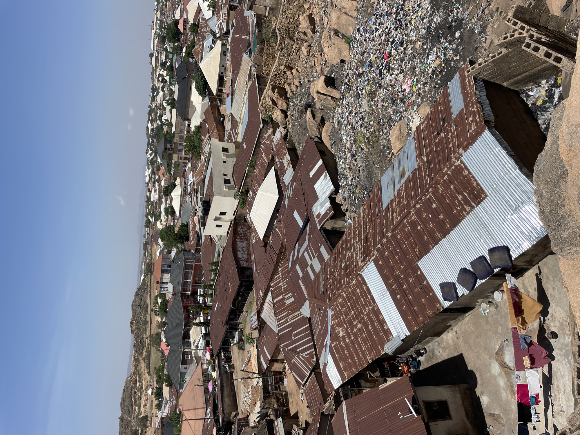 This image shows a high-angle view of a densely packed residential area in Jos, Nigeria. The roofs of the houses are primarily corrugated metal sheets, many showing signs of rust. Some roofs are partially or entirely covered with new, silver-colored sheets. The housing is closely packed with limited space between structures.
A large pile of garbage is visible, indicating the presence of waste management issues. The environment includes rocky terrain interspersed with the housing.
In the foreground, there's a concrete platform with laundry hanging to dry. Some cushions and buckets are placed on the platform. The sky is light blue with some atmospheric haze.