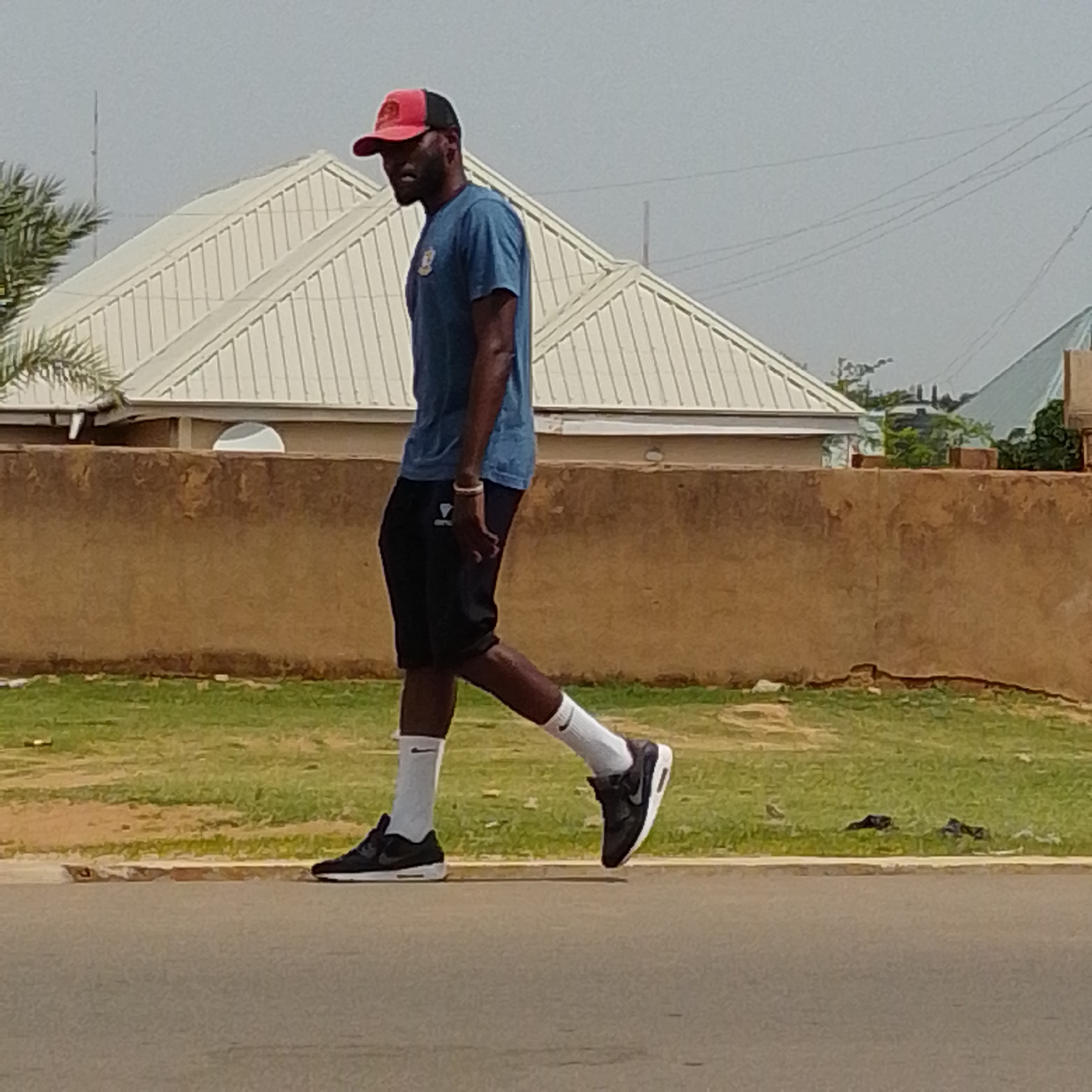 A man wearing a red and black cap, a blue t-shirt, and black shorts is walking on a paved surface. He has white socks and black sneakers. In the background, there is a brown wall, a grassy area, and a building with a white, ridged roof. The scene appears to be outdoors in a residential area, possibly in Gombe, Nigeria.