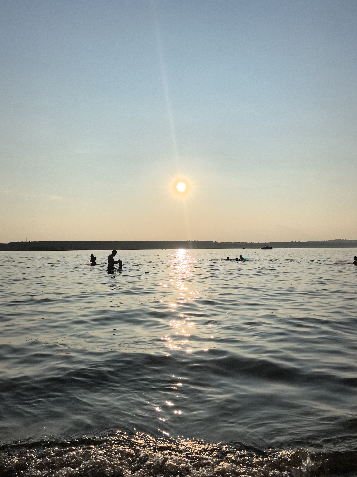 A low-angle shot captures a sunlit body of water with several individuals visible. The sun, a bright orb with lens flare, is positioned in the upper center of the frame, casting a shimmering reflection across the water's surface. The water is rippled, with small waves lapping at the foreground. In the middle ground, silhouettes of people are seen wading or swimming. One person, in the left-center, appears to be an adult with a child. Further out, other figures are visible, some appearing to swim. A sailboat is present in the distance, and the far bank of the water is lined with trees, suggesting a natural setting. The sky is clear and a pale blue, transitioning to a warmer tone near the horizon. The scene evokes a sense of summer recreation. The location is not specified to be in the city of Perm, Russia.