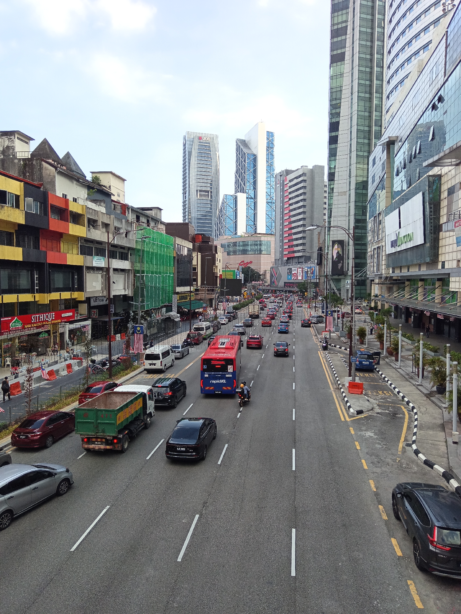 This image captures a bustling street scene in Kuala Lumpur, Malaysia, likely along Jalan Raja Laut or a nearby thoroughfare, characterized by a mix of low-rise shophouses and towering modern skyscrapers. The foreground shows a busy multi-lane road filled with traffic, including a red rapidKL bus, a green dump truck, various sedans, and a motorcycle. Pedestrians are visible on the sidewalks, and some shops with awnings are open. Flags, likely Malaysian, are displayed on some buildings and poles. The architecture displays a stark contrast between older, colorful commercial buildings on the left and sleek, glass-fronted high-rises on the right, indicative of urban development. The sky is partly cloudy. The overall impression is one of a vibrant, active urban environment.