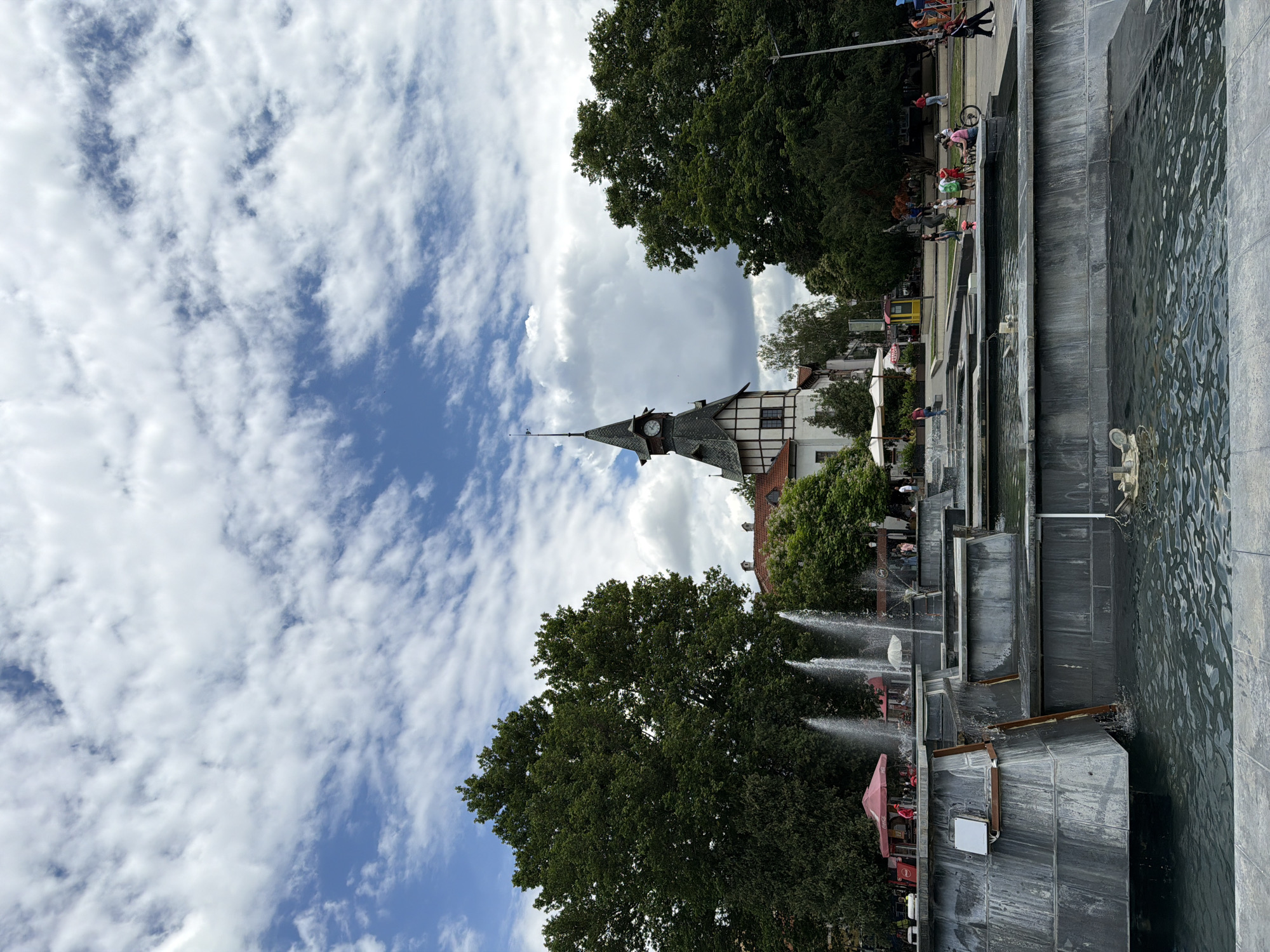 In Pazardzhik, Bulgaria, a public fountain is situated in front of a distinctive building with a clock tower. The fountain features multiple tiered levels with water spouting from some of them. The building behind the fountain is multi-storied with a steep, dark-tiled roof and a prominent clock face on its tower. Surrounding the fountain and building are lush green trees, and a clear blue sky with scattered white clouds is visible overhead. Several people are present in the scene, with some gathered around the fountain's edge, others walking on the paved area behind it, and a few riding bicycles. Red umbrellas from what appear to be outdoor seating areas are also visible near the fountain. The overall atmosphere suggests a sunny day in a lively urban park or square.