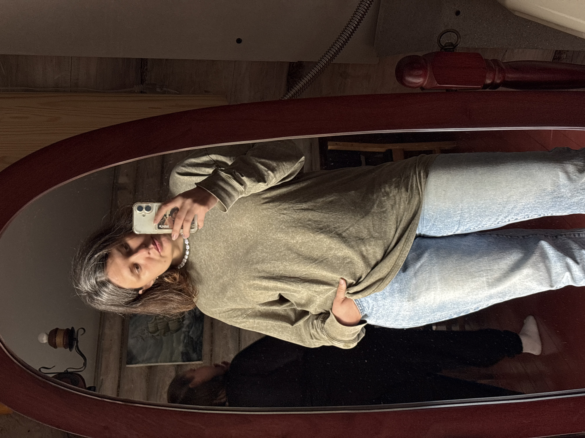 A woman is taking a mirror selfie in a dimly lit room, possibly in Костоваты, Russia. She has long, grey-streaked brown hair and is wearing a loose, olive-green long-sleeved shirt and light-wash jeans. Her right hand is in her pocket, and she is holding a smartphone in her left hand to take the photo. In the mirror's reflection, another person is visible standing further back in the room, wearing dark clothing and white socks. The room appears to be a home, with wooden walls and furniture visible. A large, ornate mirror with a dark red frame dominates the foreground.