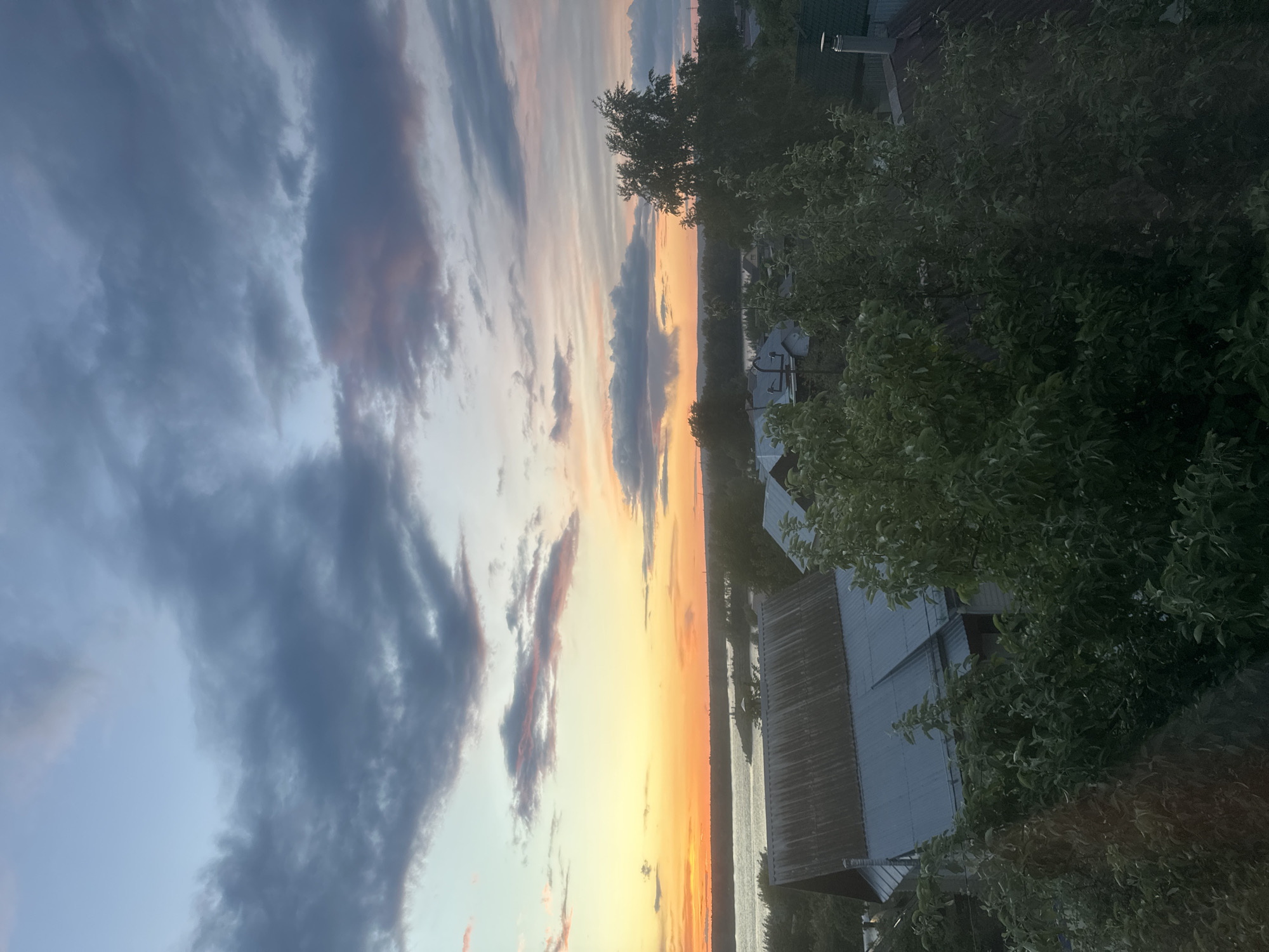 The image shows a landscape at sunset in Chaikovsky, Russia. The sky is filled with a mixture of dark, heavy clouds and bright orange and yellow hues from the setting sun. Below the sky, there is a body of water, possibly a river or a lake, reflecting the light. On the right side of the frame, there are trees and the roofs of buildings, adding a sense of location.