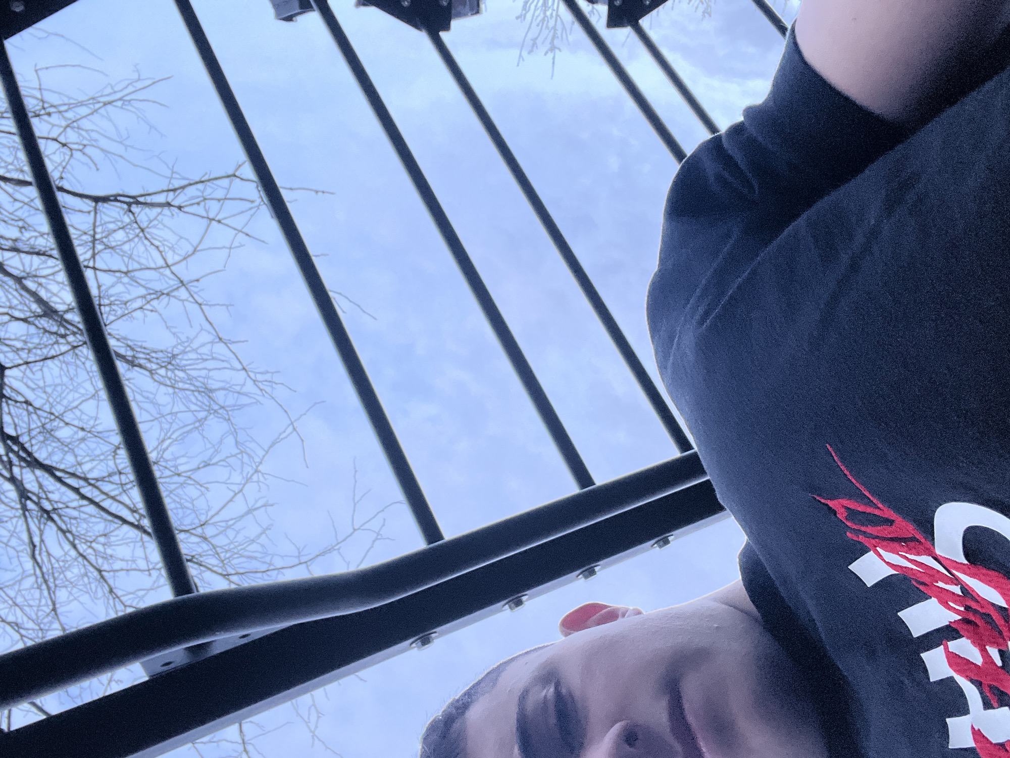 The image is an upward-angle shot of a person lying underneath a black metal structure, possibly a staircase or similar architectural element. The person is wearing a dark-colored shirt with a design featuring white and red elements. In the background, a pale blue sky with some cloud cover is visible between the metal bars. Bare tree branches extend into the frame from the left side. The location is inferred to be Nova Zagora, Bulgaria, although this cannot be definitively verified from the image.