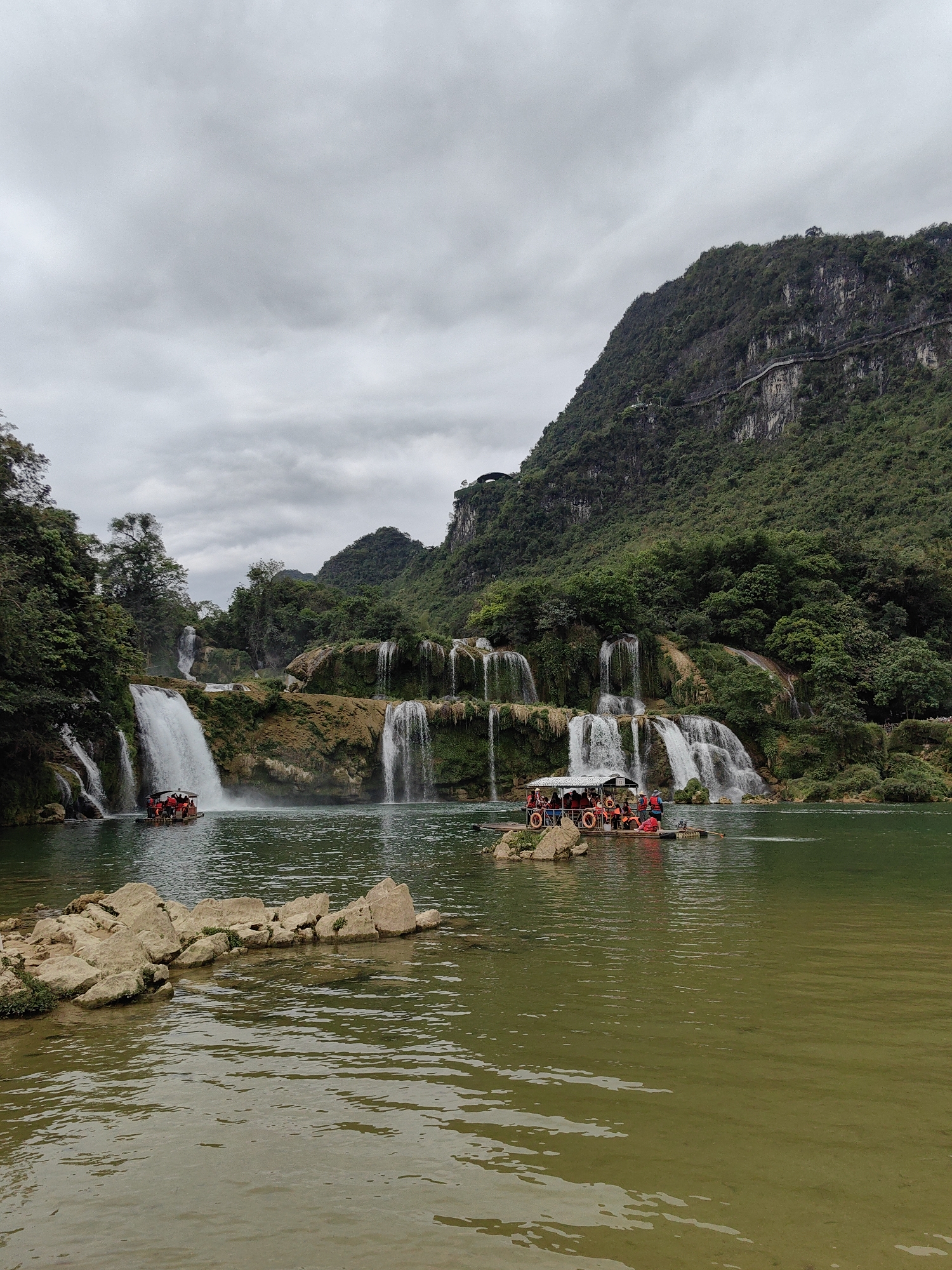 The image shows the Detian Waterfalls in Daxin County, Guangxi, China. Several waterfalls cascade down a rocky, vegetated hillside into a green river. Two rafts are visible in the river, each carrying a group of people wearing life jackets. A rocky outcropping is visible in the foreground. A mountain covered in trees rises in the background, with a path carved along its side. The sky is overcast and grey.