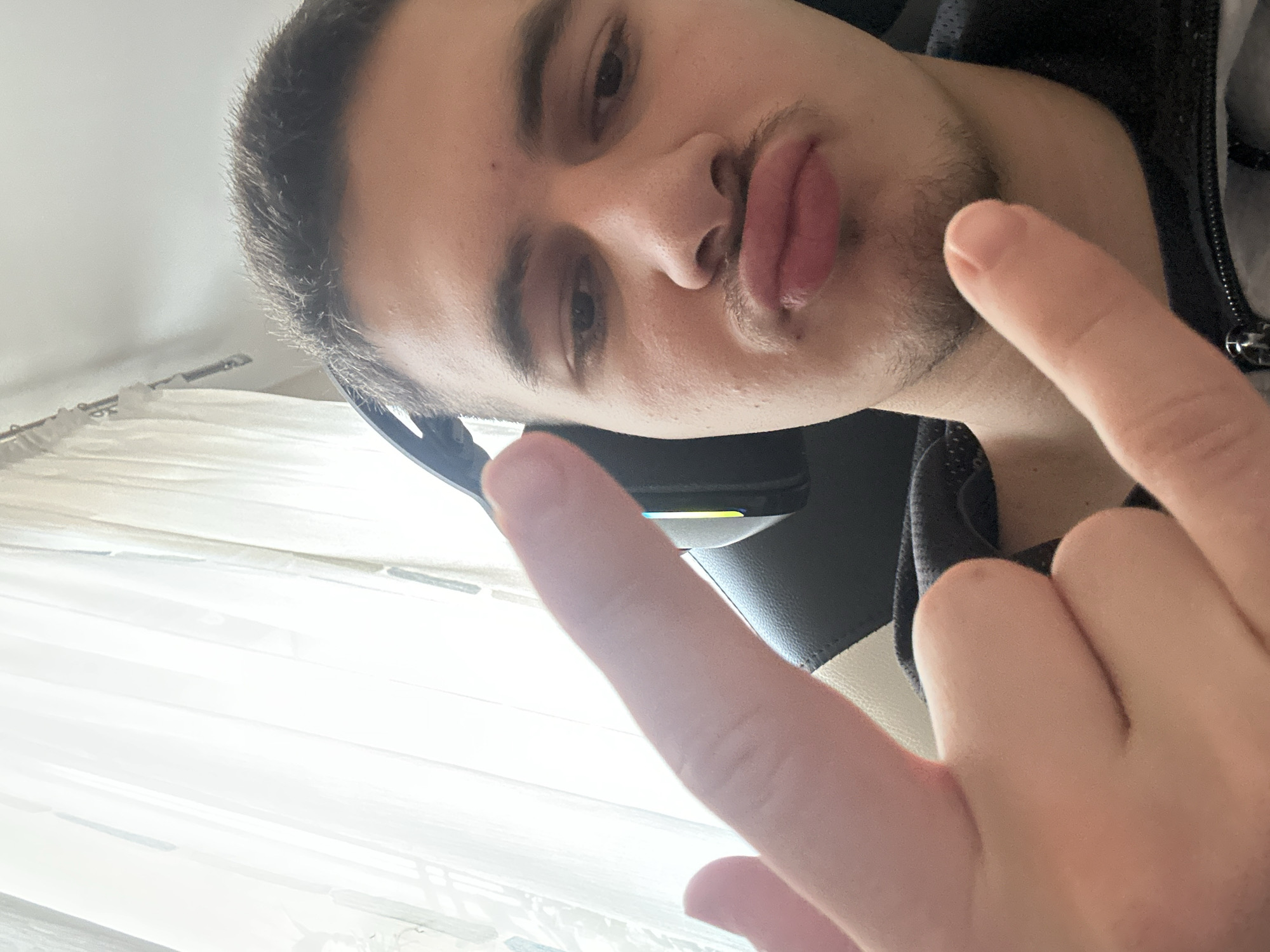 A selfie of a young man with short dark hair, light skin, and stubble is shown. He is wearing headphones and a dark-colored jacket. He has his lips pursed slightly and is holding his hand up with his index and pinky fingers extended, making a "rock on" or "devil horns" gesture. The background appears to be indoors near a window with light filtering through the curtains. The location in Sofia, Bulgaria, cannot be determined based on the visual information provided.