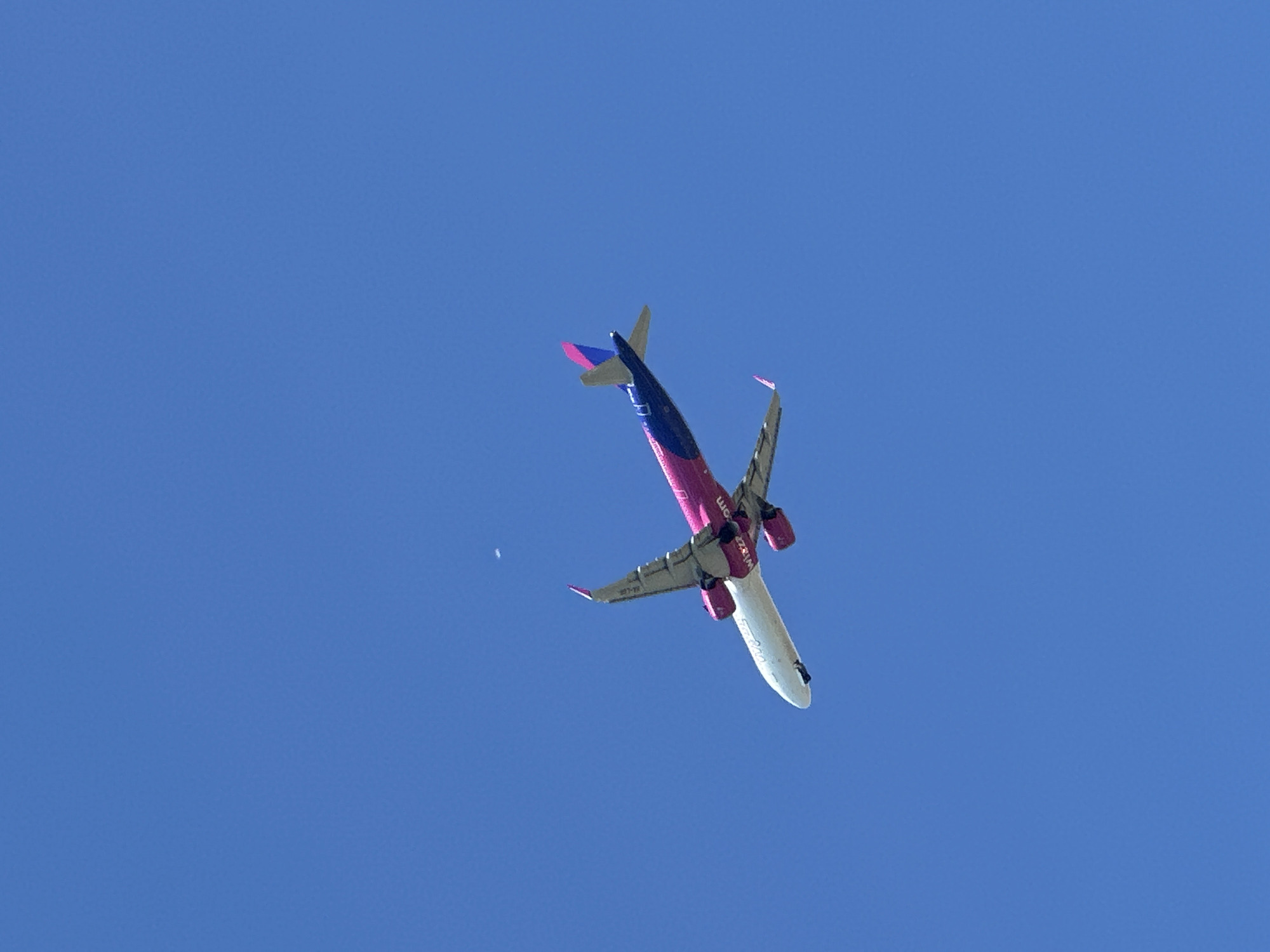 A low-angle shot shows a Wizz Air airplane against a clear blue sky. The airplane is pink and white. It is oriented diagonally from the bottom right to the upper left of the frame. The tail has a blue and pink design. The photograph was taken from Varna, Bulgaria.
