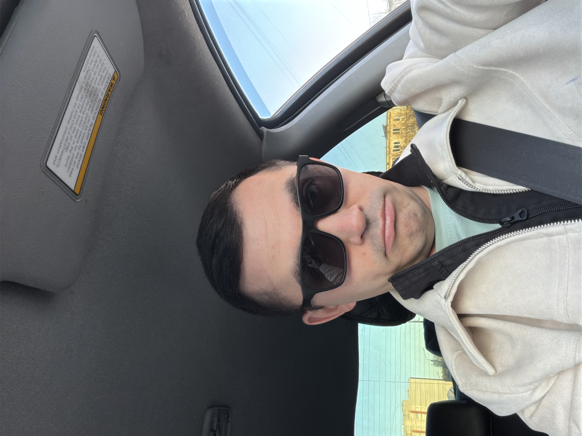 Here is a description of the image:
A man is in the driver's seat of a car, wearing sunglasses and a light-colored jacket. He is looking at the camera. The car has a sunroof, and the sky is visible through it. A warning label is visible on the car's sun visor. Outside the car windows, a building is visible. The city location is not specified.